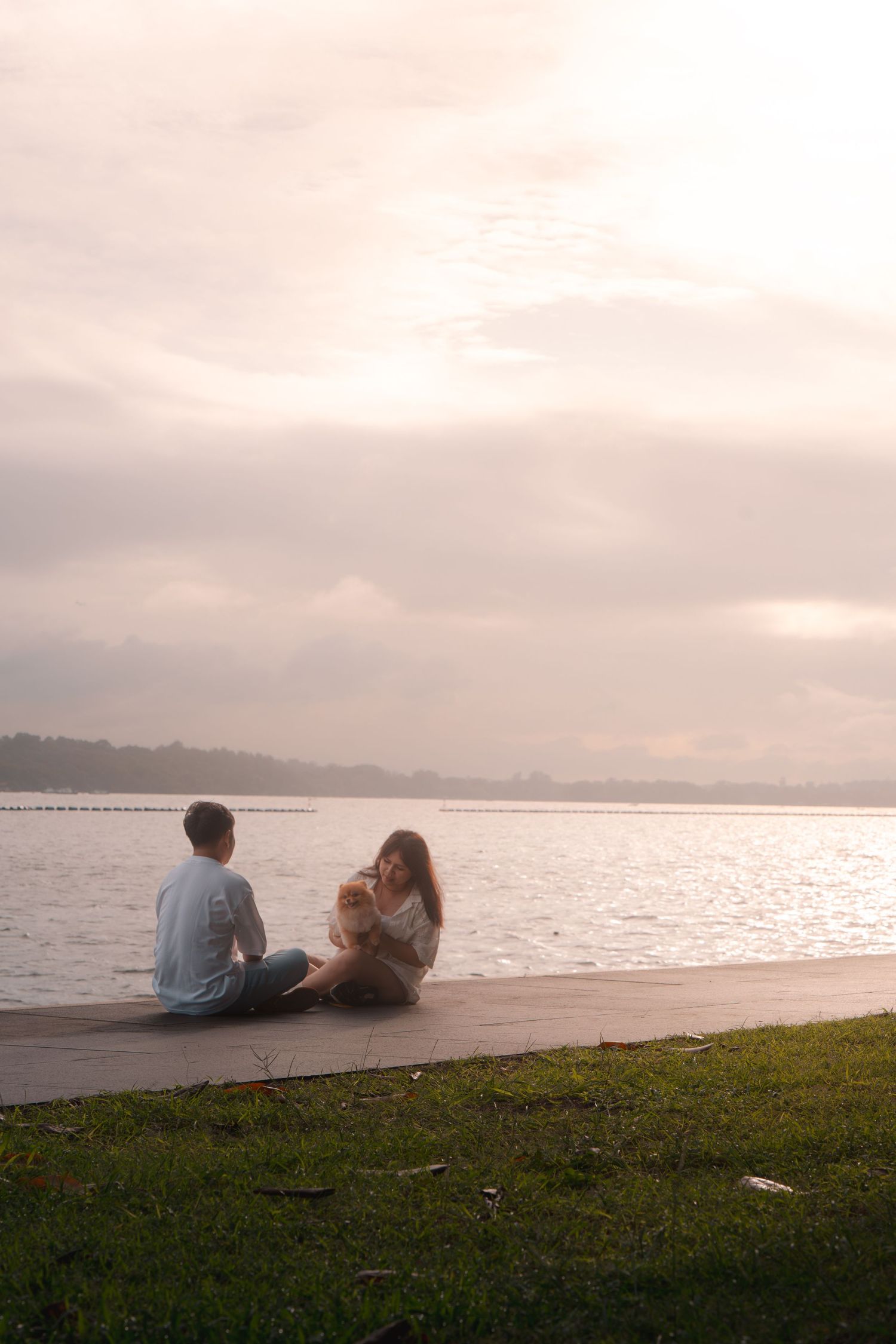 Couple sitting together on a waterfront promenade at sunset with a small dog, enjoying a peaceful view of the calm lake.