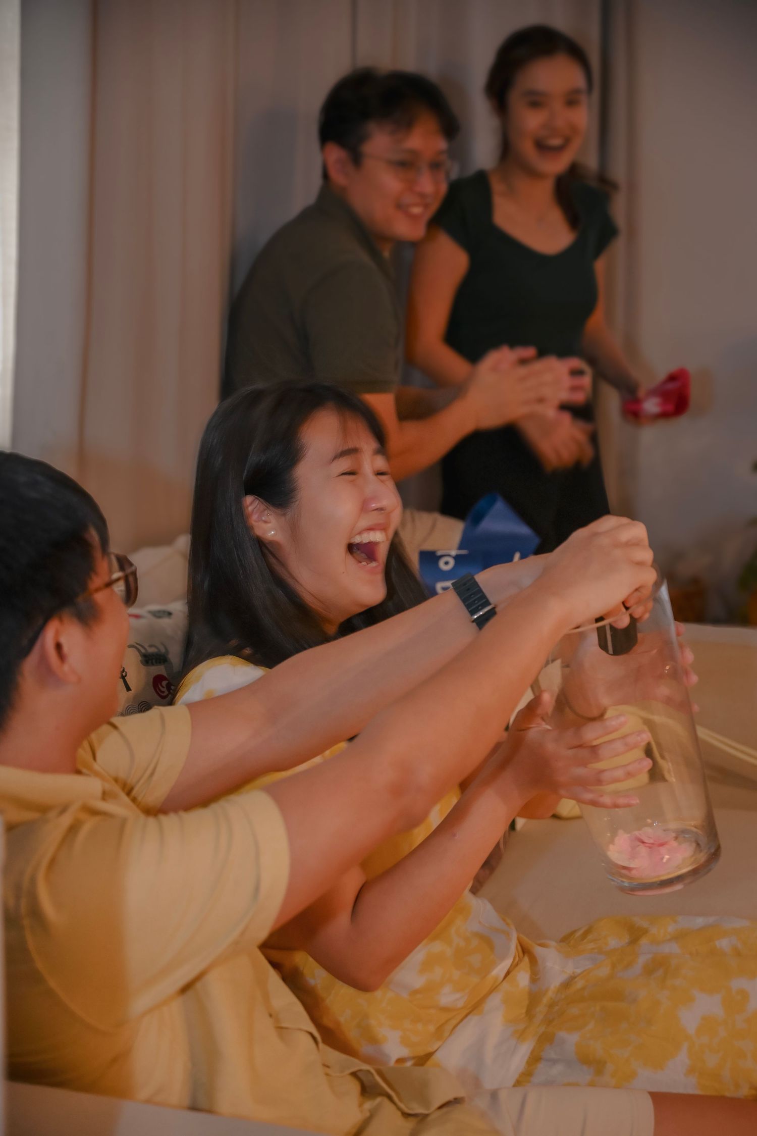 Group of Asian friends laughing and toasting drinks at a lively indoor gathering party.