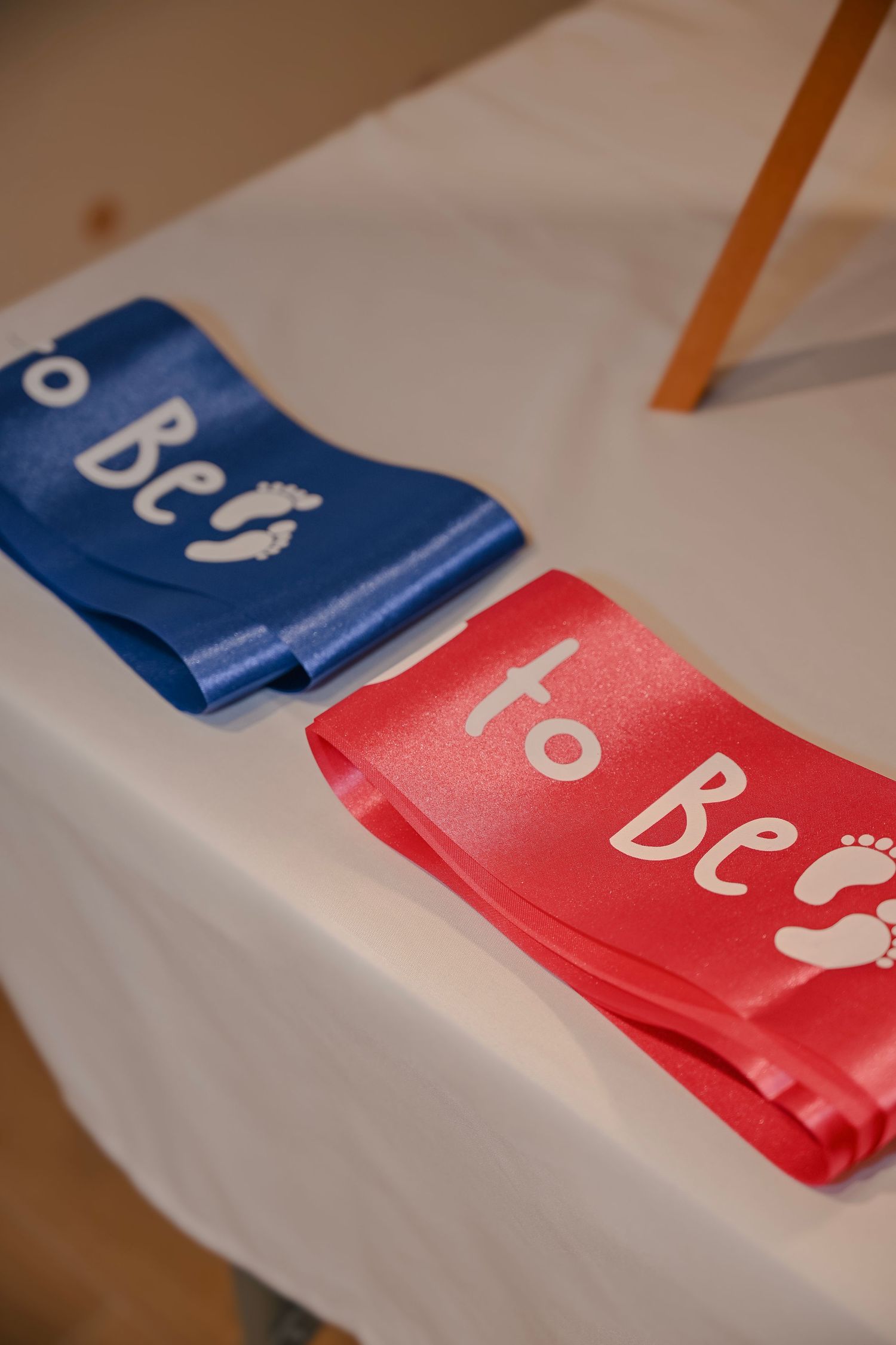 Blue and pink satin 'Mom to Be' and 'Dad to Be' sashes laid on a white table for a gender reveal party.
