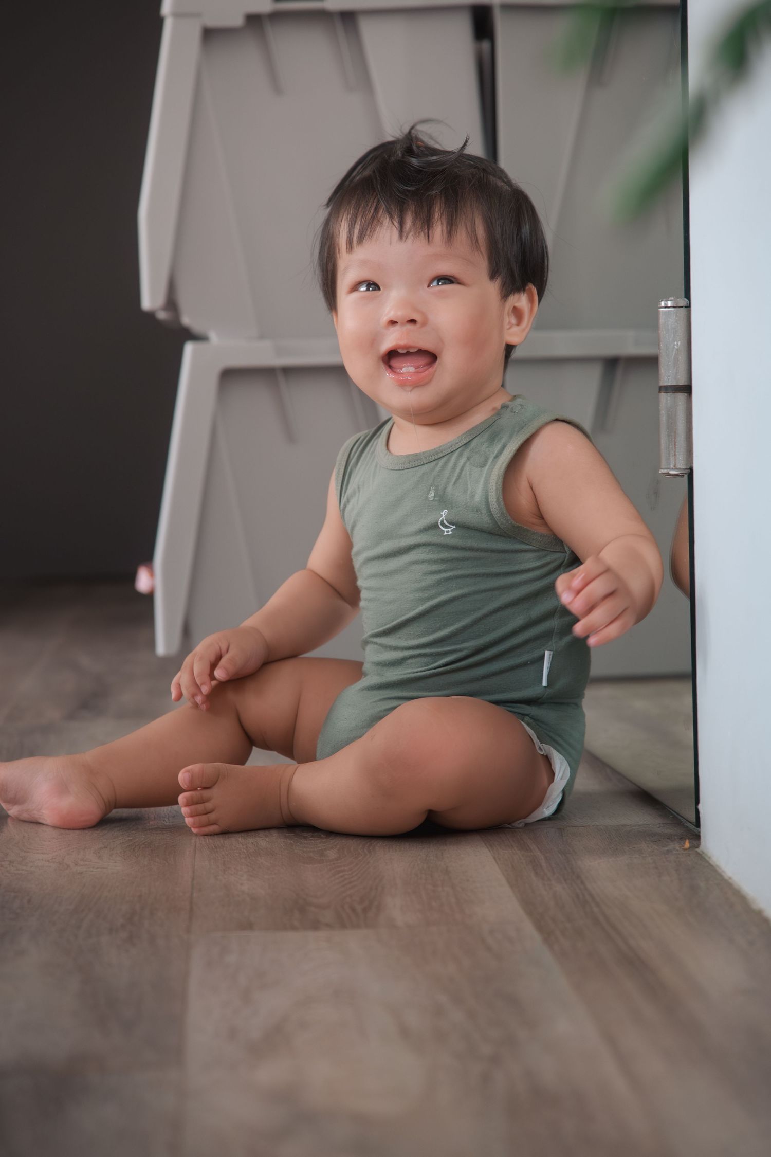 A happy baby in a green tank top sits on a hardwood floor near a white wall.