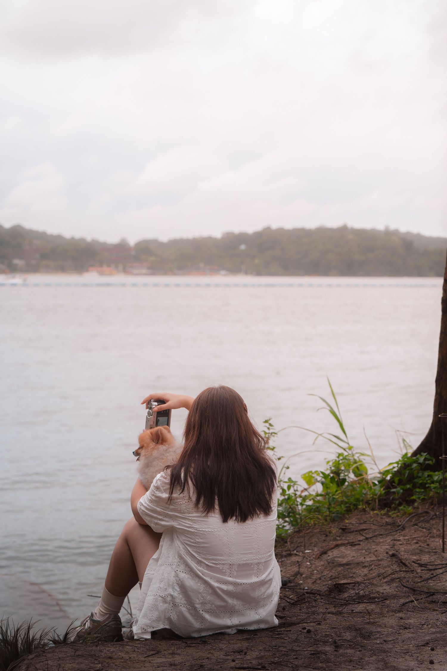 Woman in white dress sitting by a calm lake shore, holding a small dog while gazing at the water on a cloudy day.