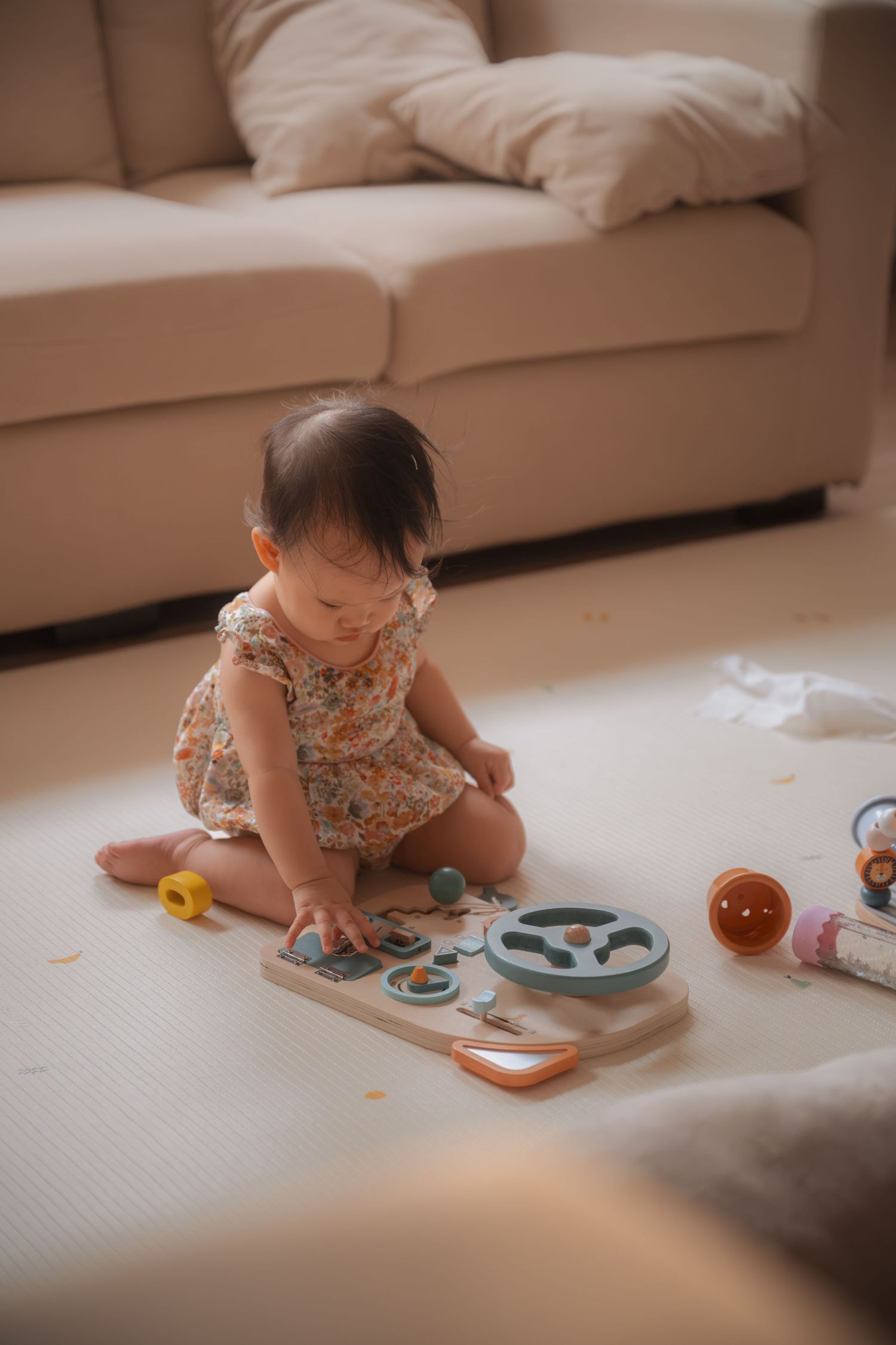 Baby girl in floral romper playing with wooden puzzle toy on floor near beige sofa.