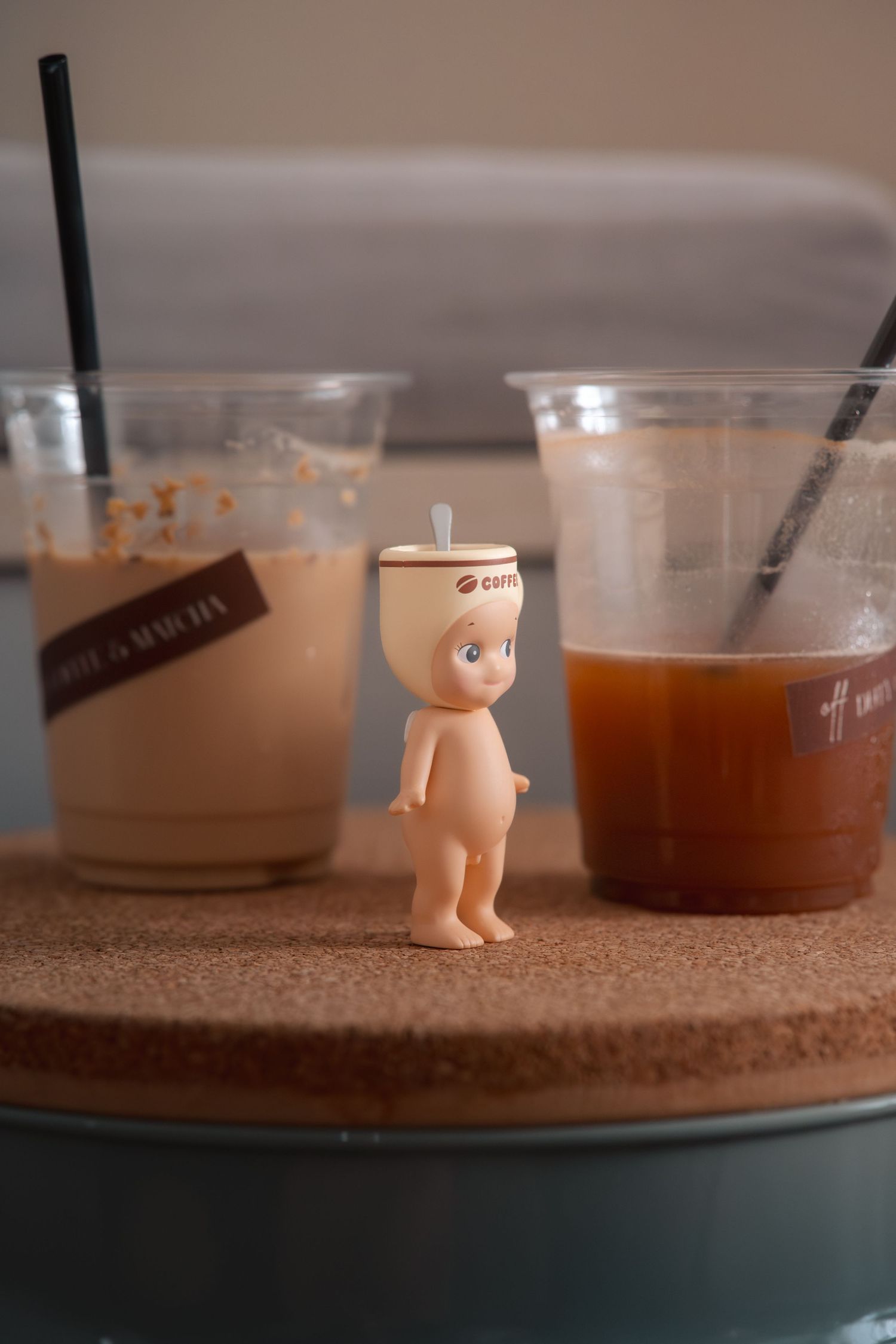 A small toy figure stands next to clear plastic cups containing brown liquid on a cork surface.