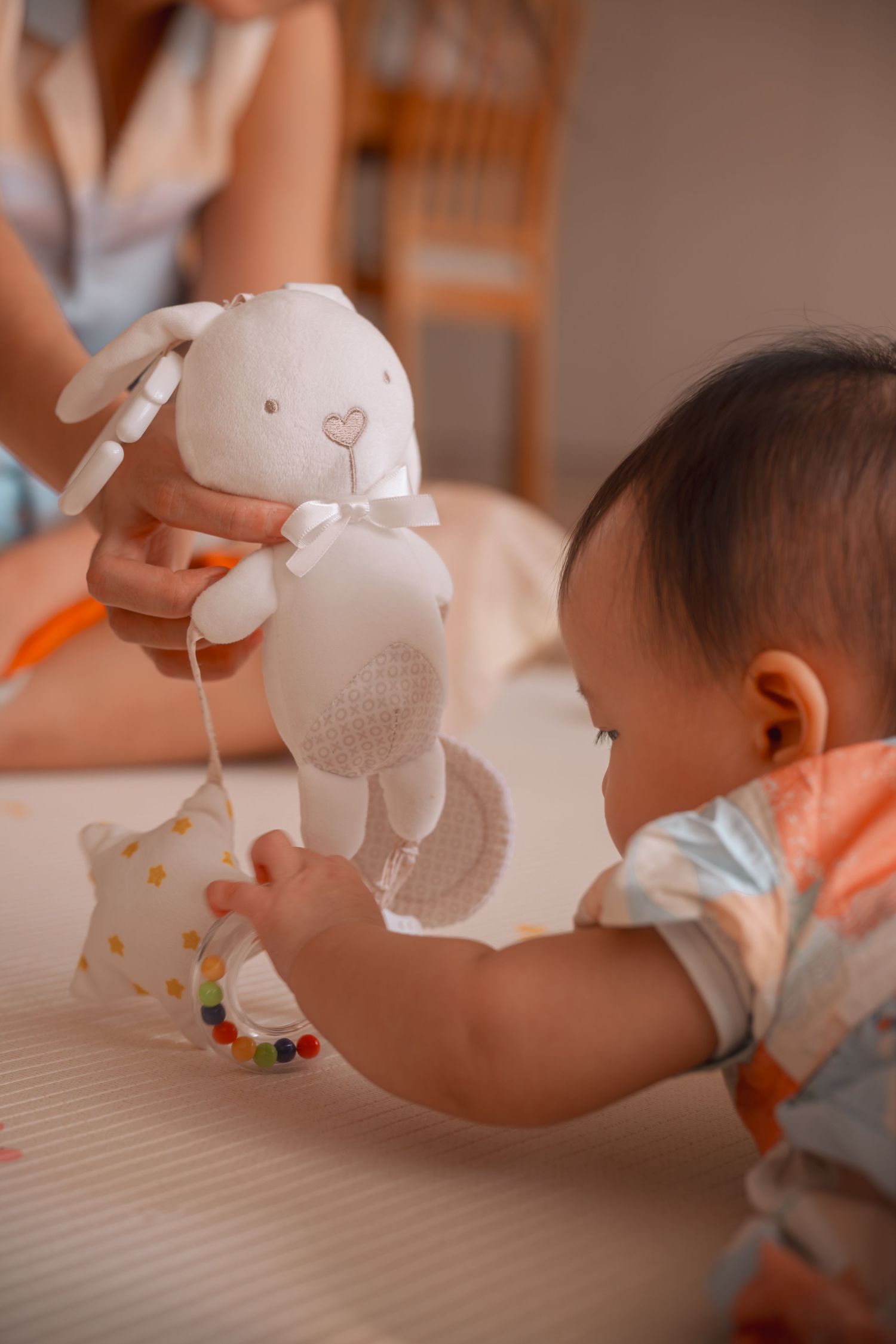 Baby reaching for a white bunny stuffed animal toy held by an adult on a soft play mat.