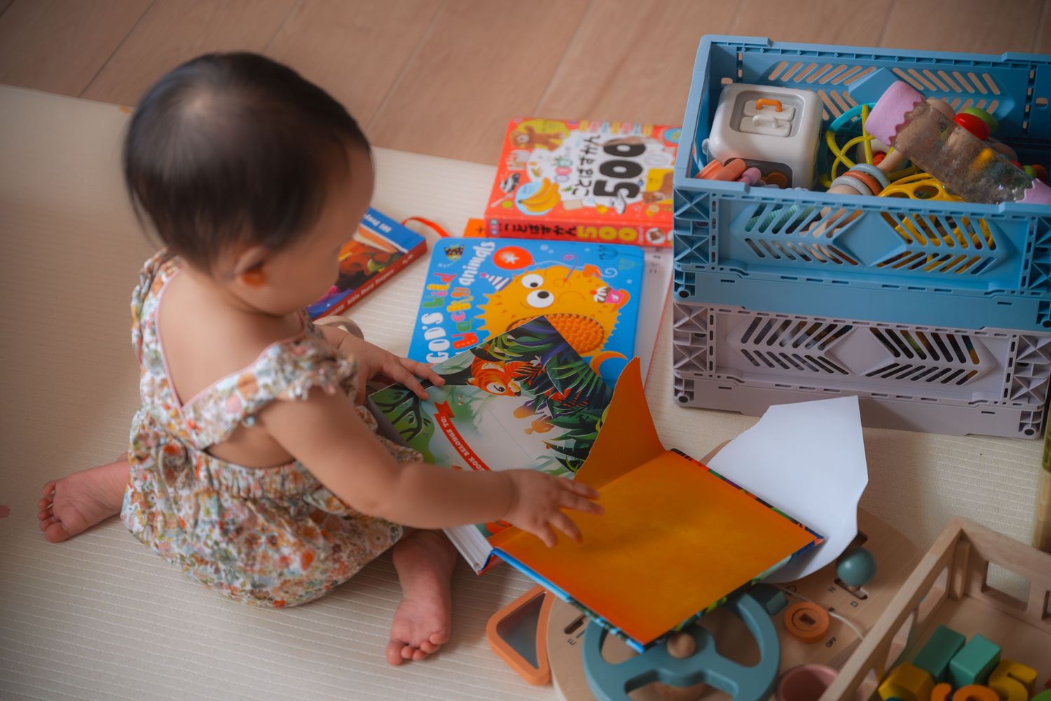 A baby girl sits on the floor exploring colorful children's books and toys from a blue storage bin.
