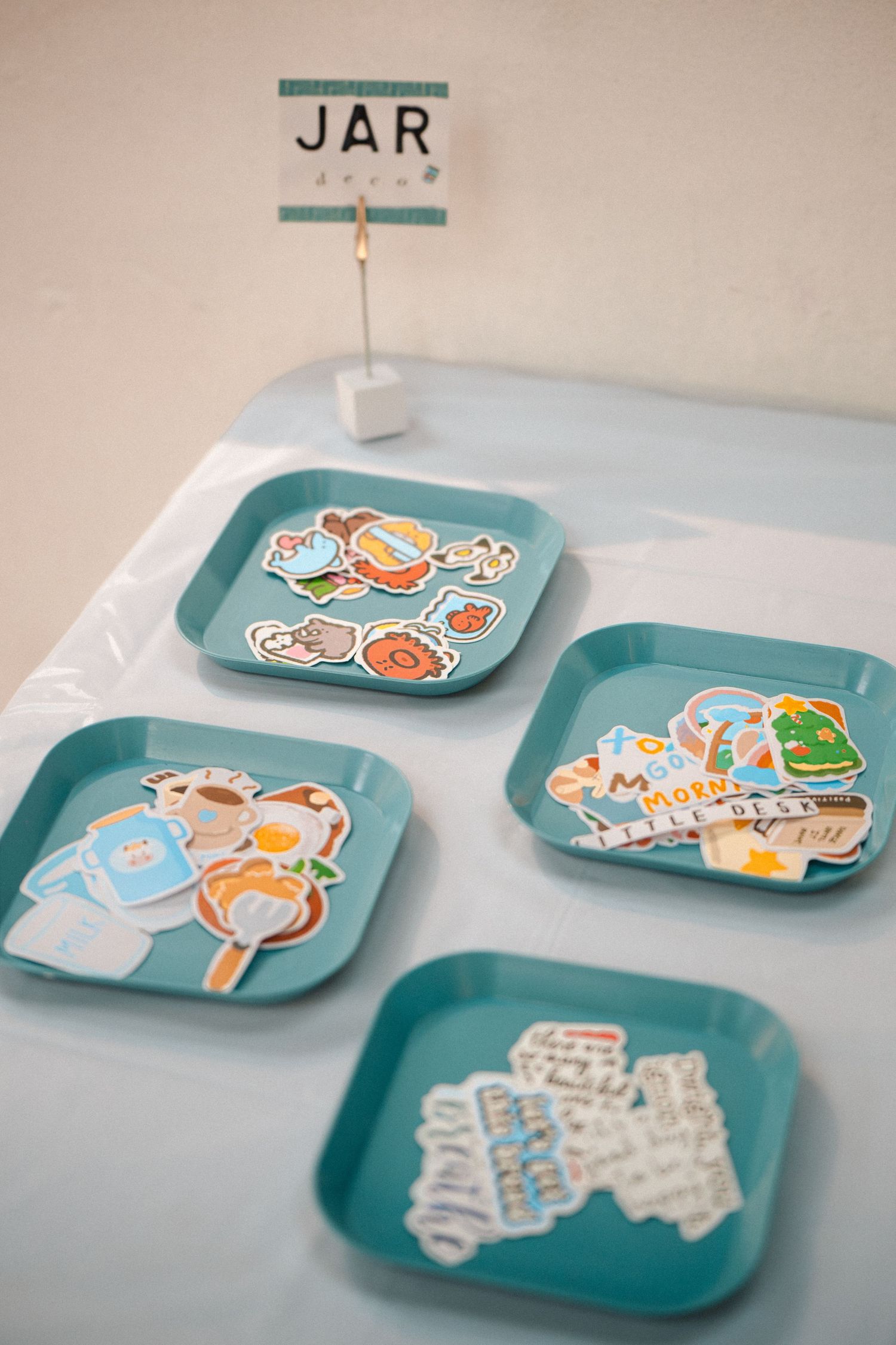Teal square trays displaying colorful stickers and paper cutouts on a blue tablecloth, with a JAR sign holder.