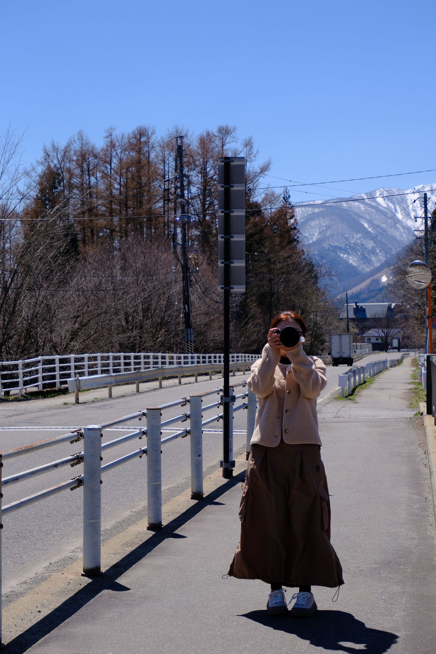 Woman photographing scenic mountain landscape on a sunny day, standing on a bridge path with snow-capped peaks and blue sky.