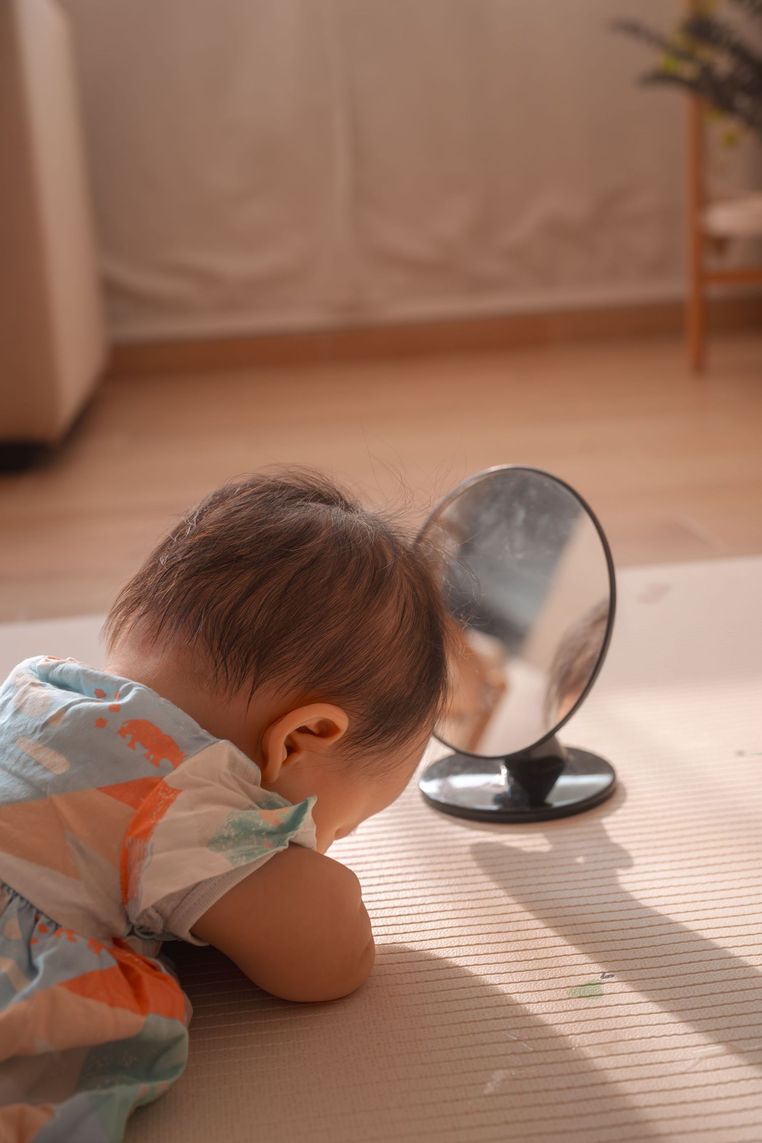 Baby in dinosaur onesie curiously looking at reflection in small round mirror on floor indoors.
