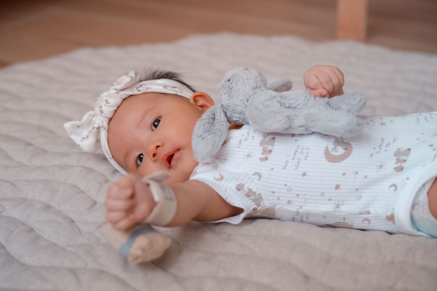 A baby wearing a white floral outfit lies on a gray blanket while looking at the camera.