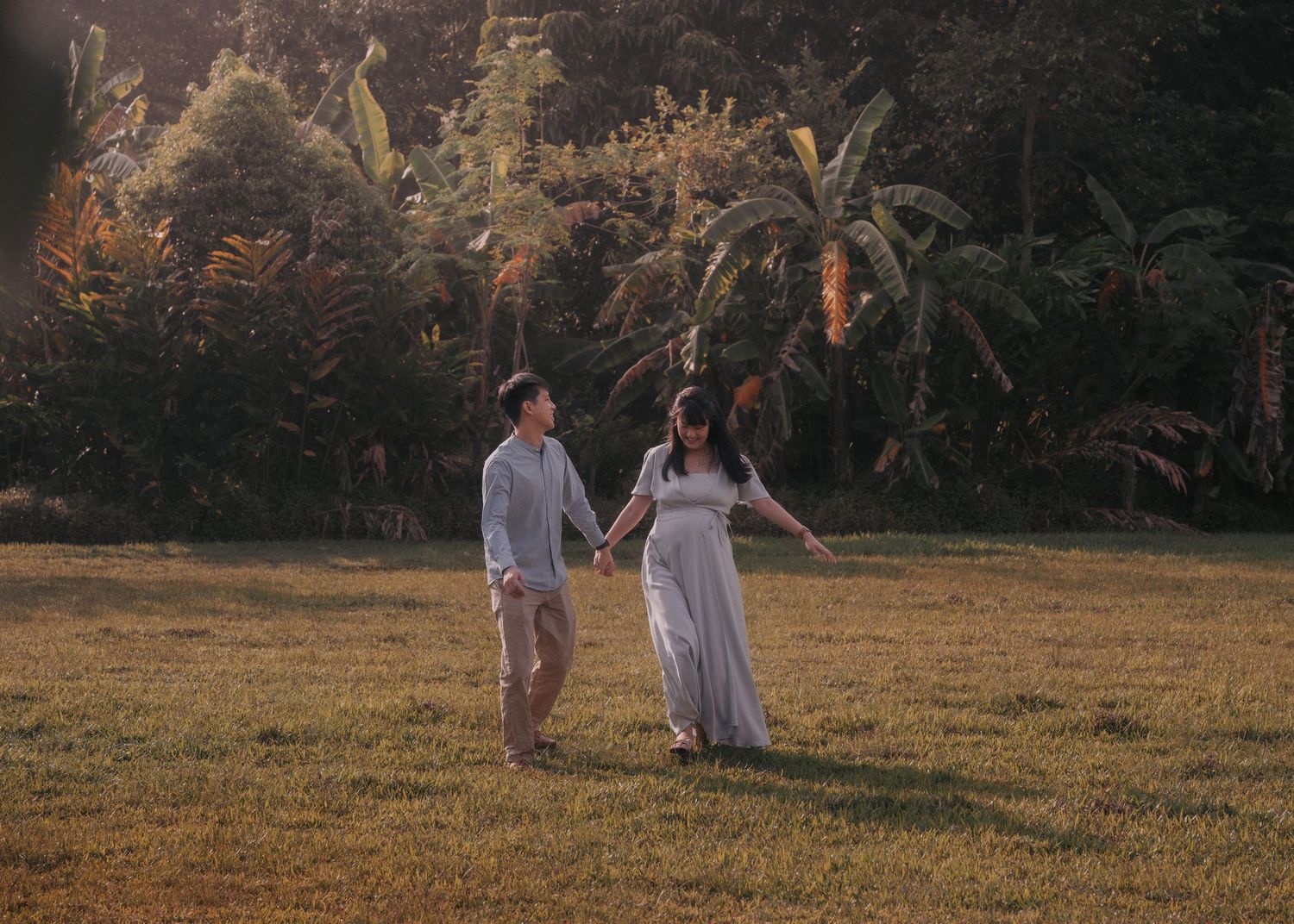 Couple holding hands walking on lush green lawn surrounded by tropical trees in warm golden sunlight.
