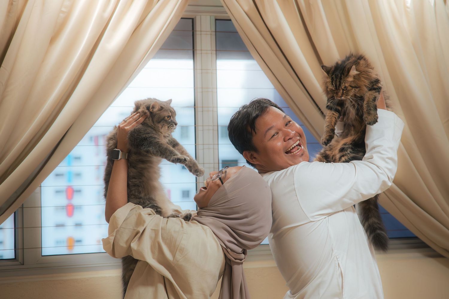 Happy man laughing while holding two fluffy cats near curtained windows in a bright, cheerful indoor setting.