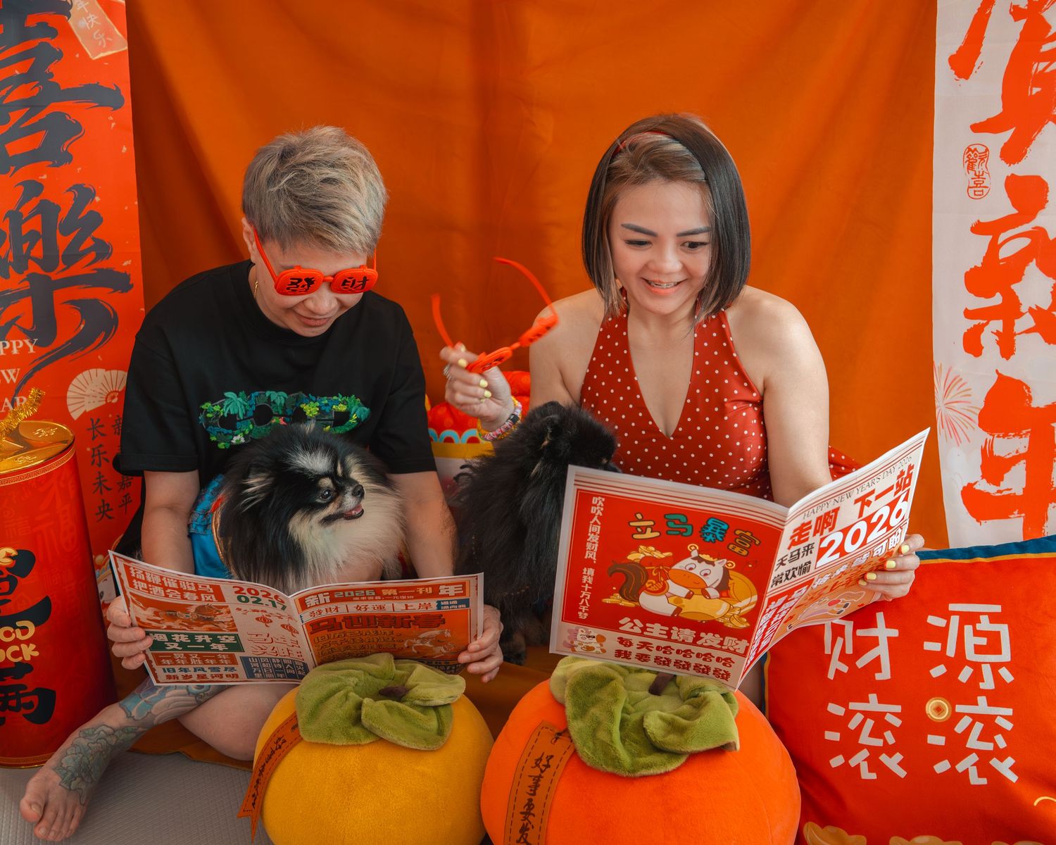 Two people reading red Chinese New Year booklets with a dog, surrounded by orange decorations and persimmons.