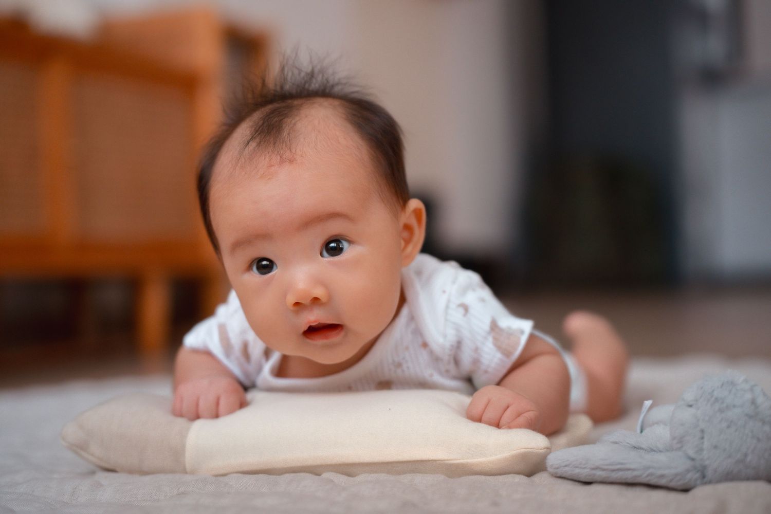 Baby in white onesie lying on soft carpet with pillow while doing tummy time at home.