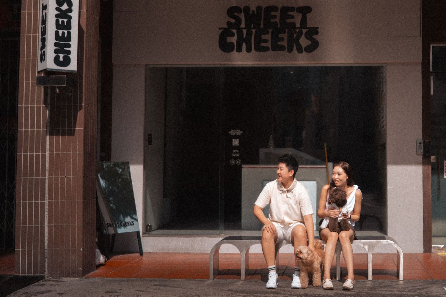 Two people sit on chairs outside a storefront with 'Sweet Cheeks' signage in Singapore.