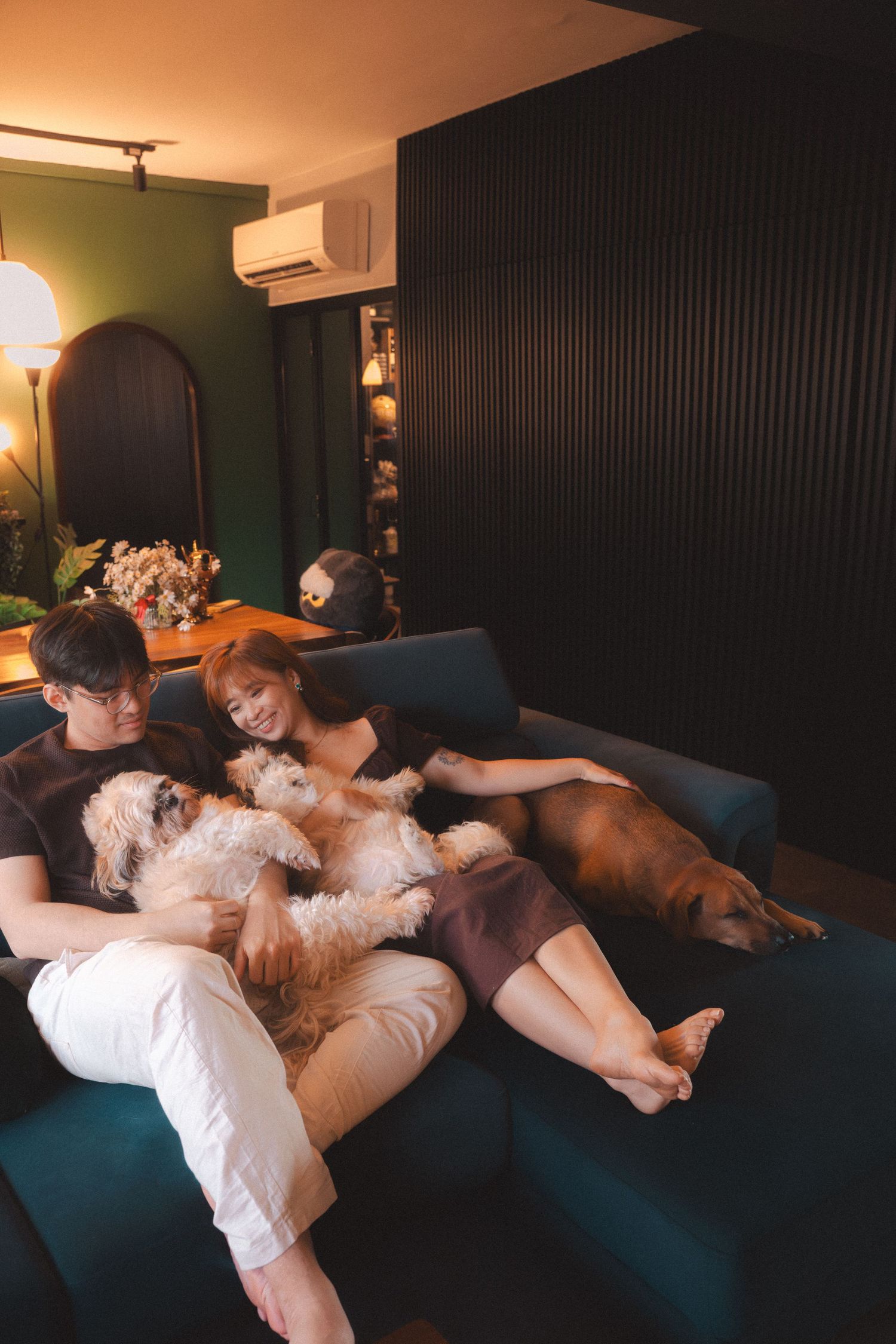 Three people and two dogs relax together on a dark blue couch in a dimly lit room with warm lighting.