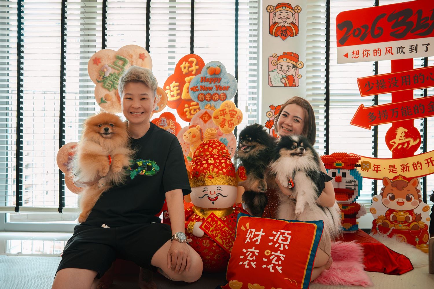 Two people holding Pomeranian dogs pose with colorful Chinese New Year decorations and festive red ornaments indoors.