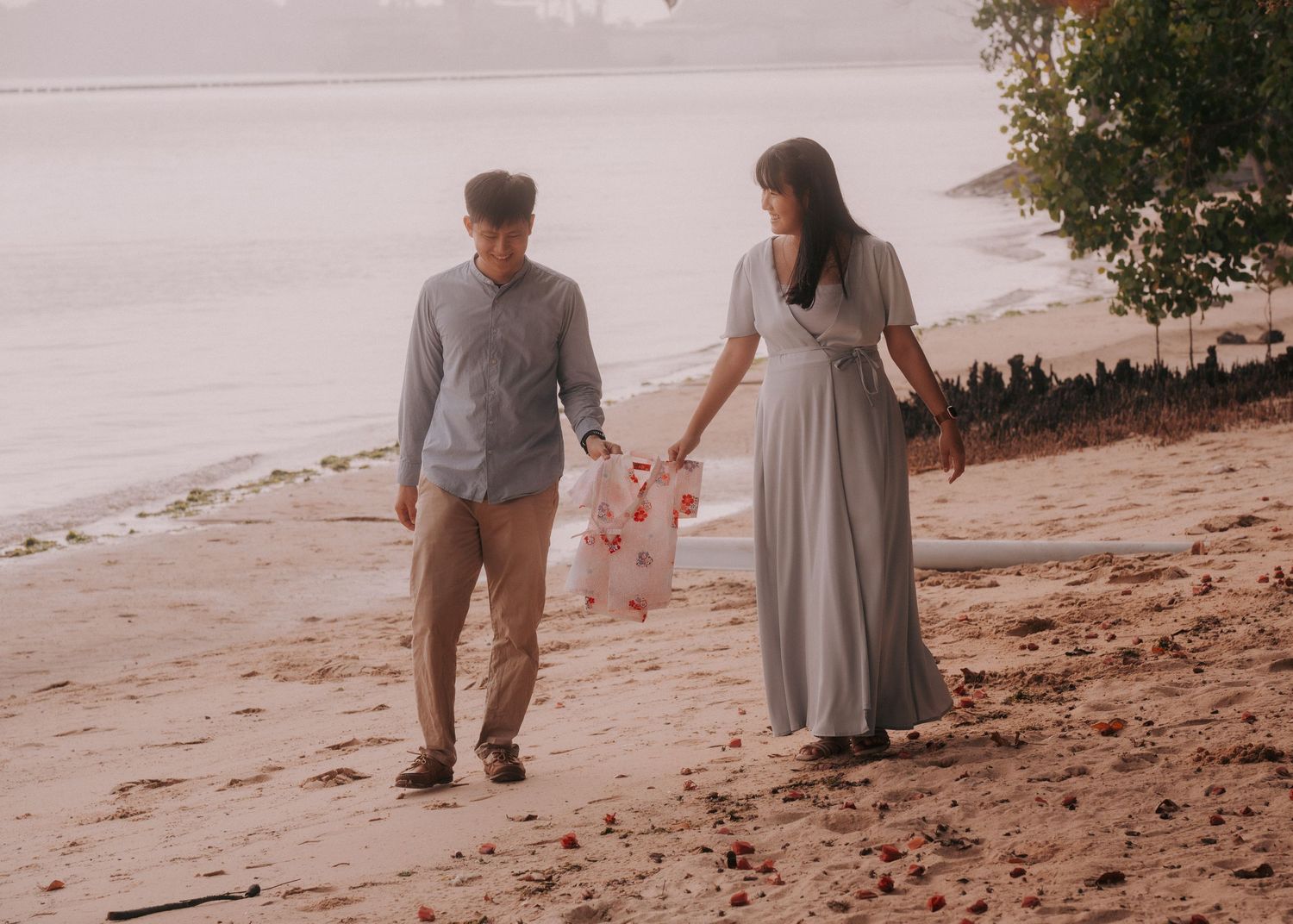 Couple walking hand in hand on a sandy beach, man in gray shirt and woman in flowing light blue dress.