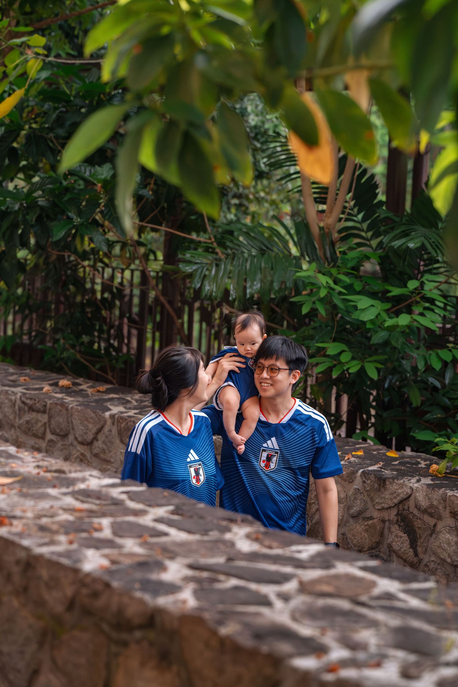 A young Asian family in matching blue Japan Adidas jerseys holds their baby outdoors near a stone wall with lush greenery.
