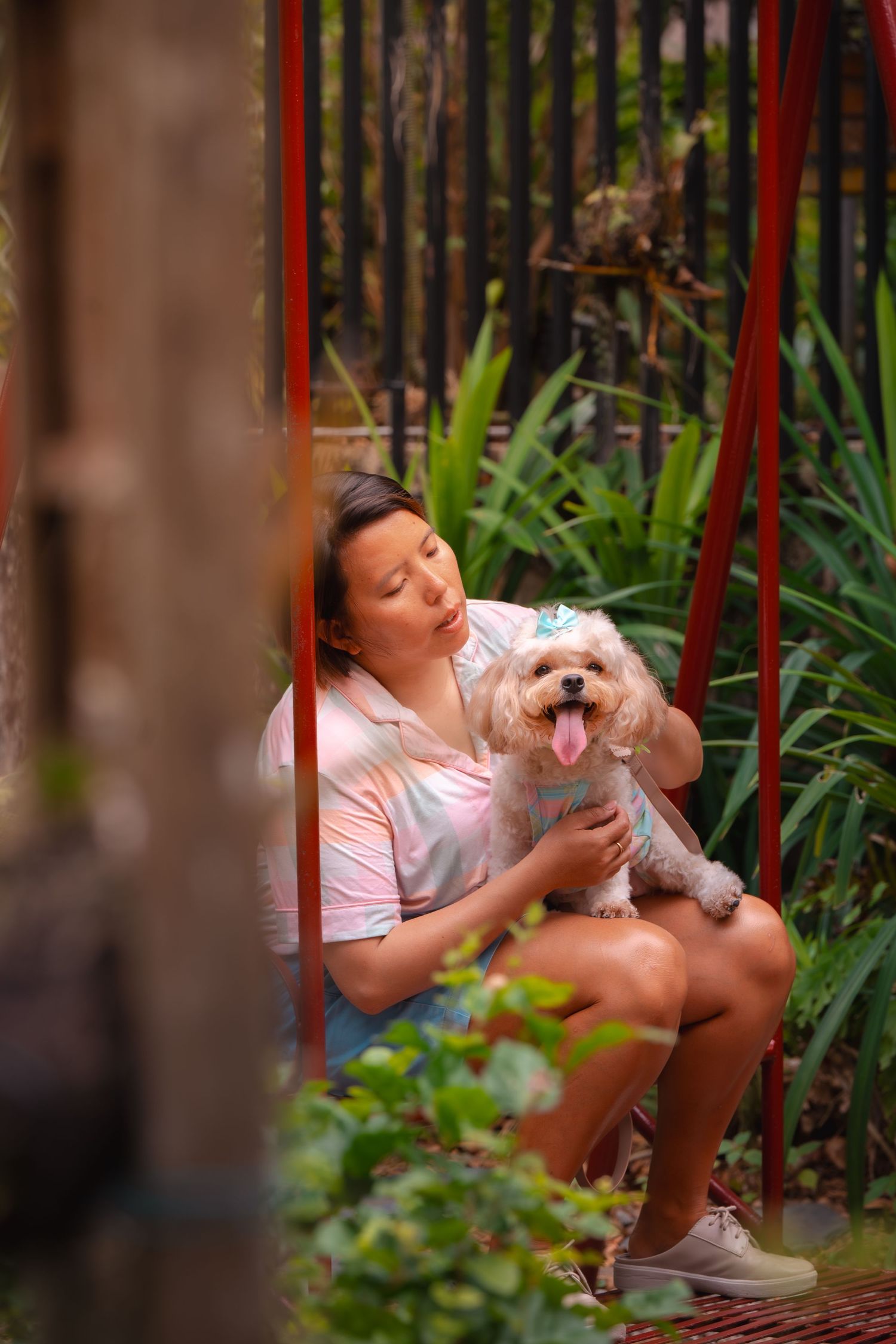 A small white dog sits on a red swing in a garden setting.