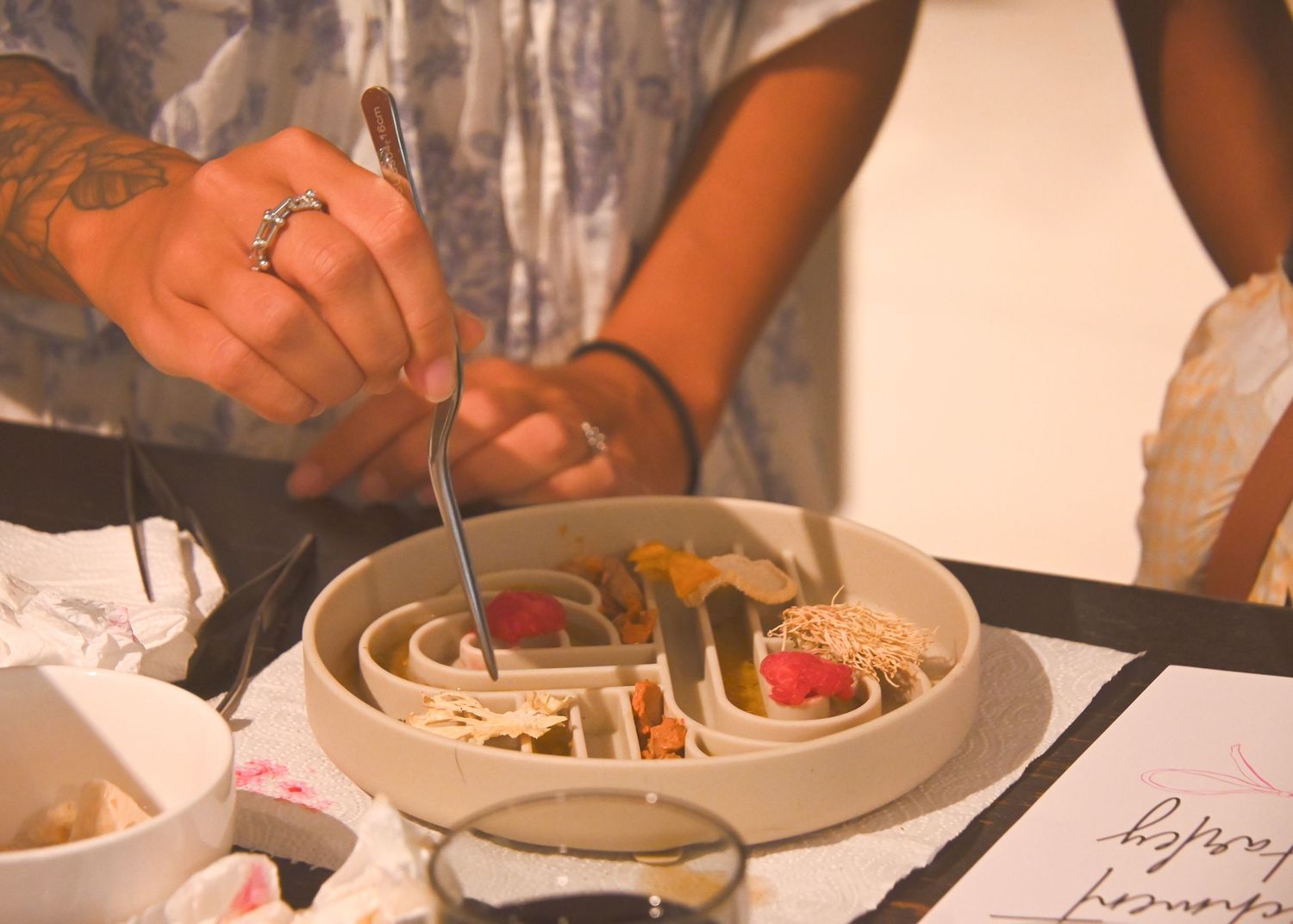Tattooed hand holding fork over elegant ceramic dish with colorful gourmet food at a fine dining restaurant table.