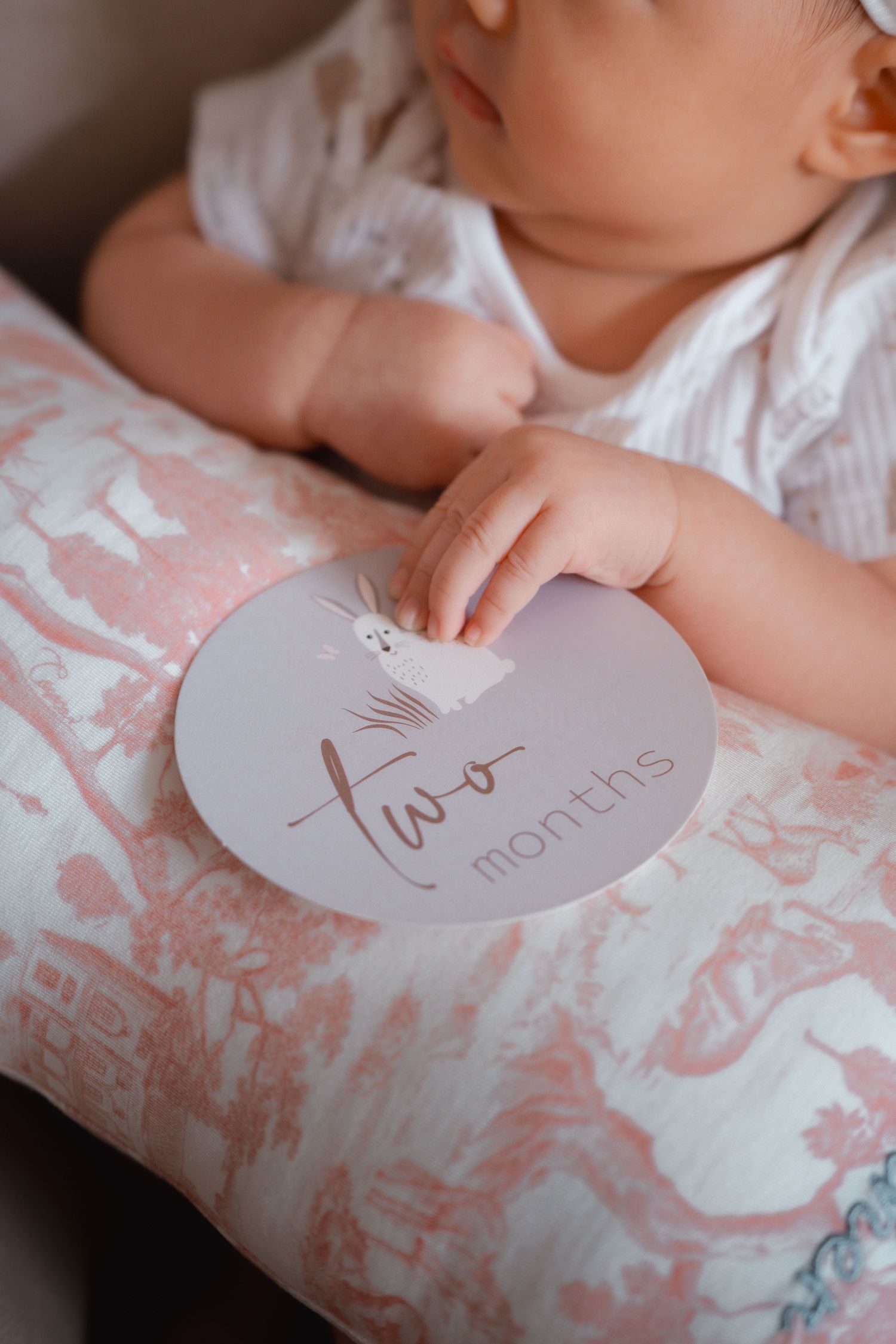 A baby's hand holds a round gray milestone card marking two months while resting on a pink and white patterned pillow.