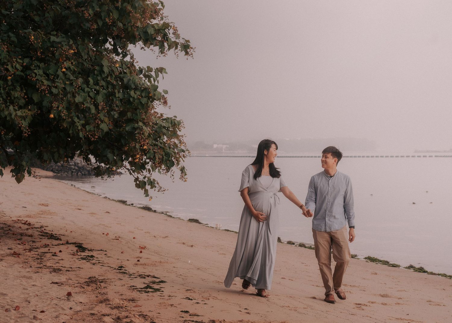 A couple strolls together on a beach with trees in the background during sunset.
