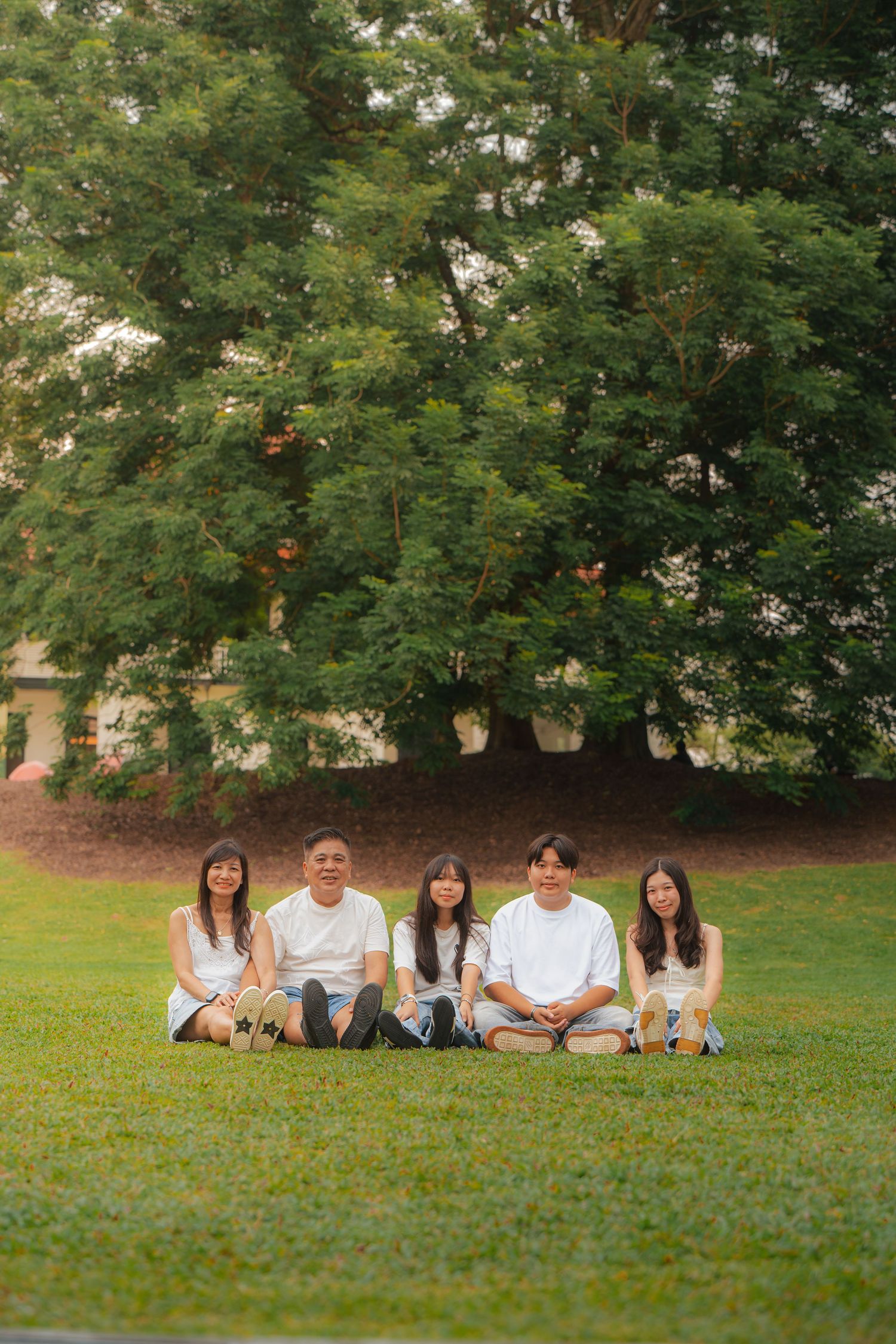 Five young adults in white shirts sitting on green grass in a park with large trees in the background.