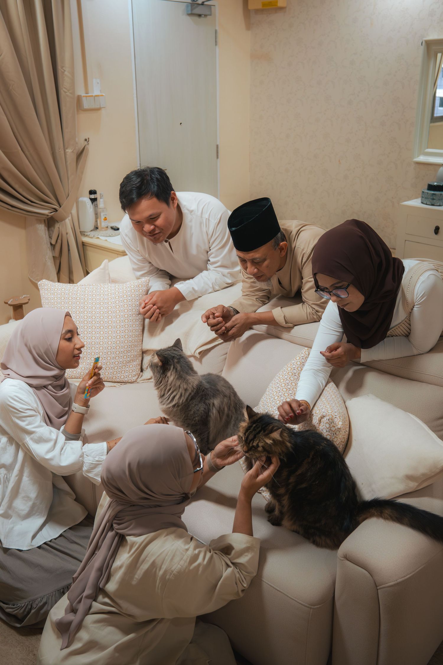 Muslim family in traditional attire gathered on sofa, petting cats together in a cozy living room.