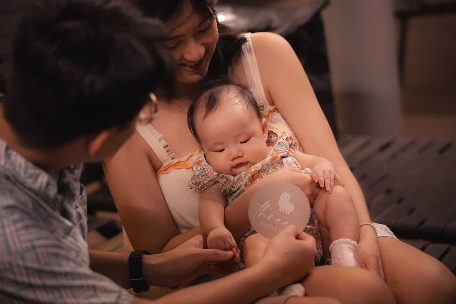 A baby in a floral outfit rests between two adults on a bed in a warm-toned indoor setting.