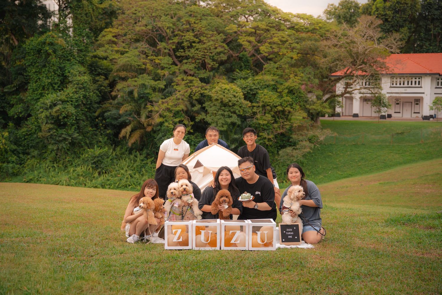 Family group photo with dogs on green lawn, decorative letter blocks spelling ZEZE, trees and white building in background.