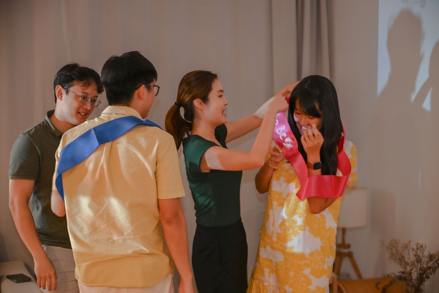 Group of people at an indoor celebration, woman placing a colorful accessory on another woman's head.