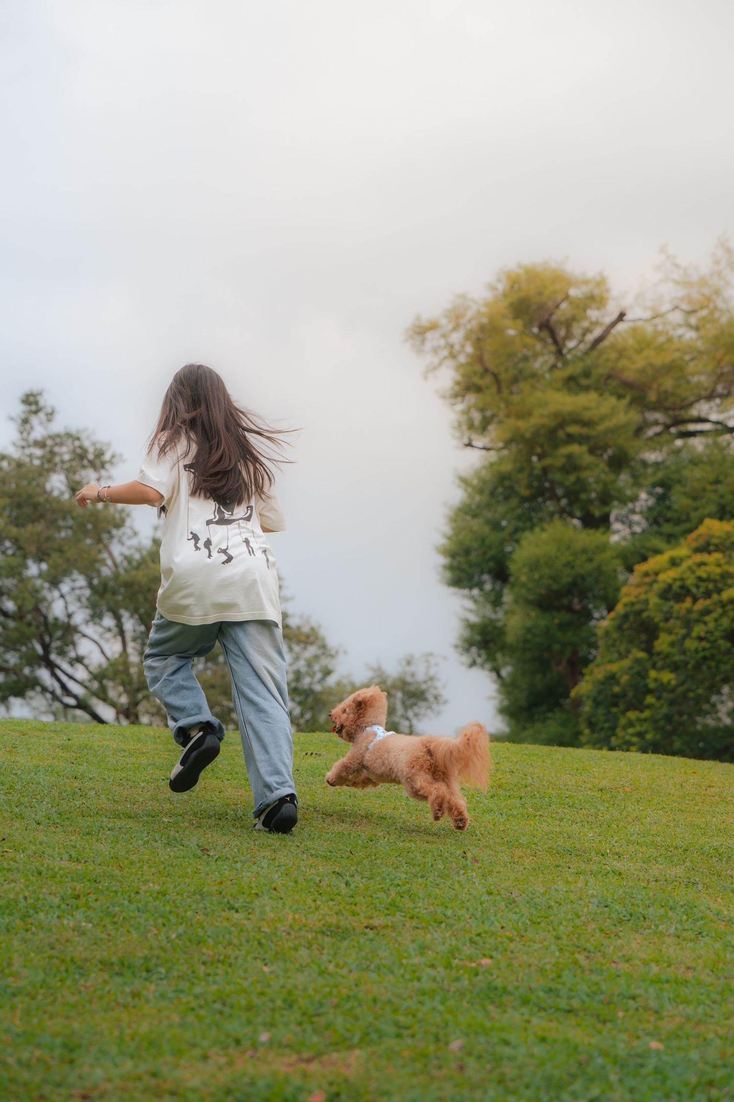 A girl runs playfully with a fluffy golden dog on a lush green hillside, trees and cloudy sky in the background.