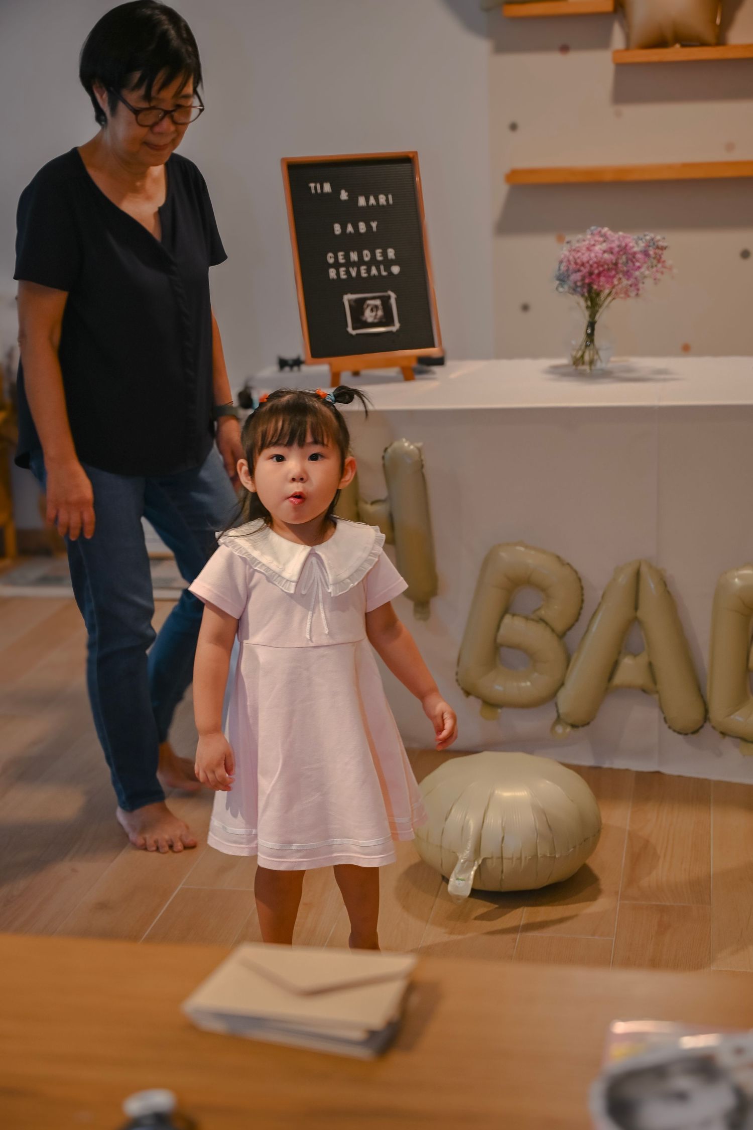 A small child wearing a white collared dress stands near balloon decorations spelling 'BABY'.