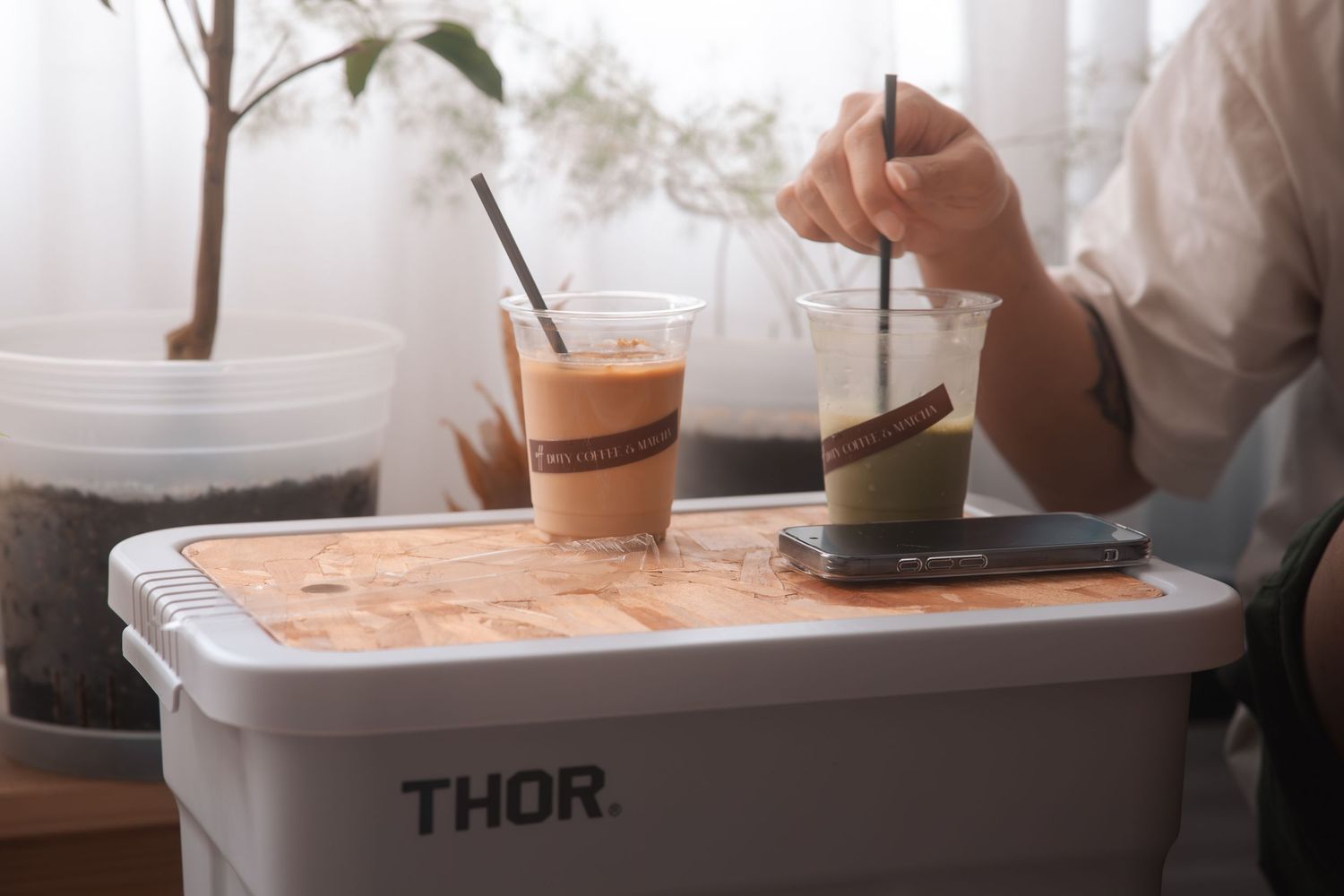 Two iced coffee drinks on a Thor cooler box, with a person stirring one of the beverages indoors.
