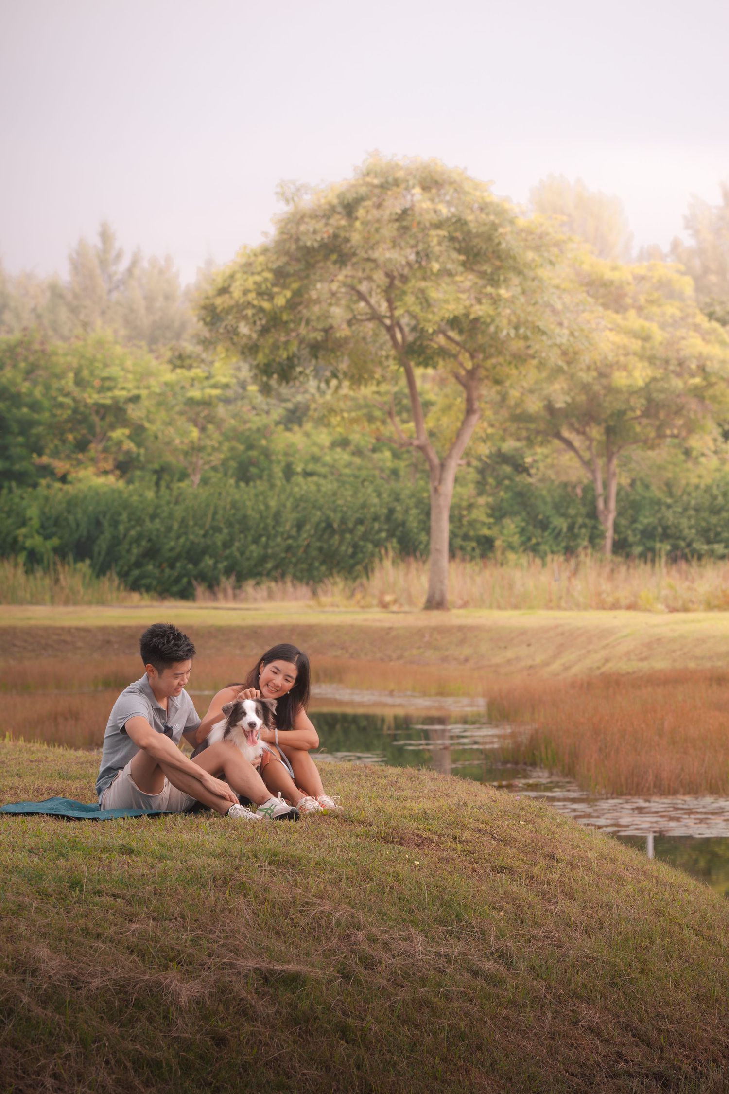 Two young people sit on grass by a pond with a small dog, surrounded by lush green trees in a serene park setting.