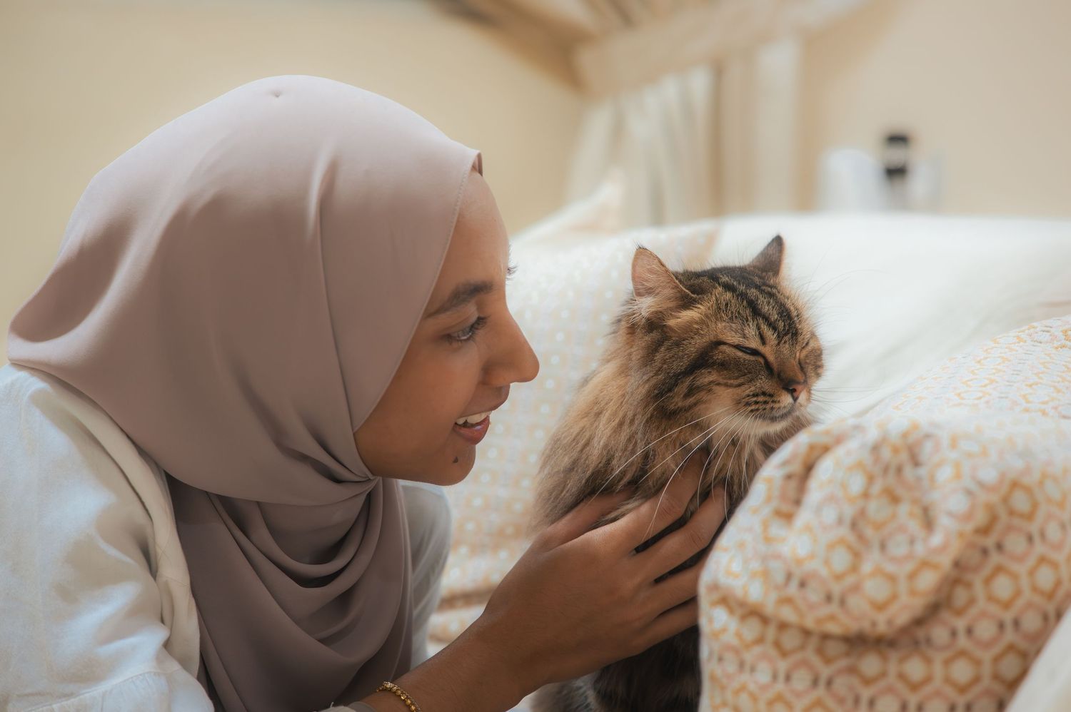 A Muslim woman in a hijab gently pets a fluffy cat while lying on a bed, sharing a tender moment.