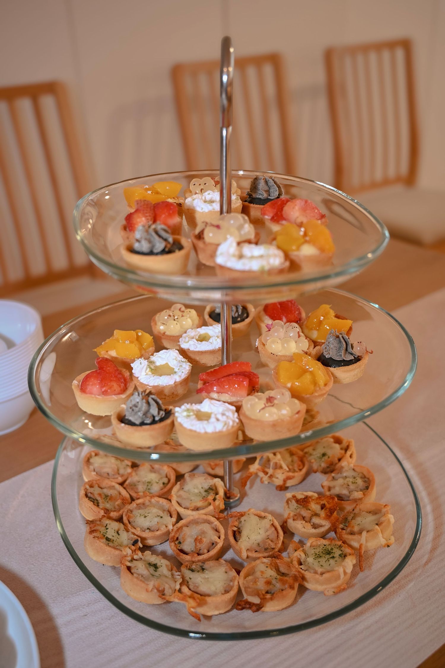 Three-tier glass serving stand filled with assorted mini tarts and savory appetizers on a wooden table.