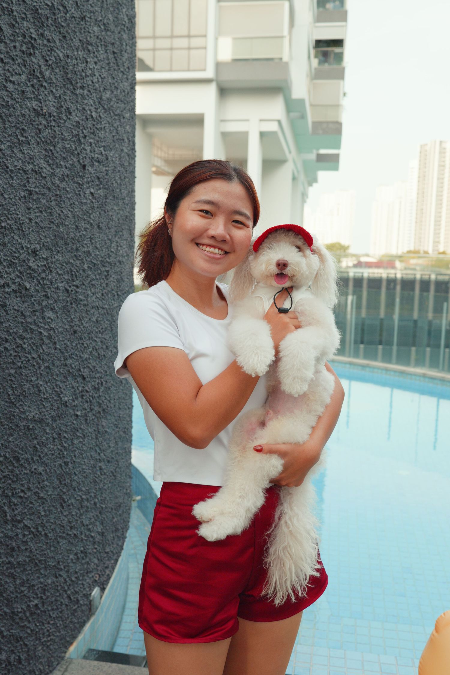 Smiling Asian woman holding a fluffy white dog wearing a red cap near a rooftop pool.