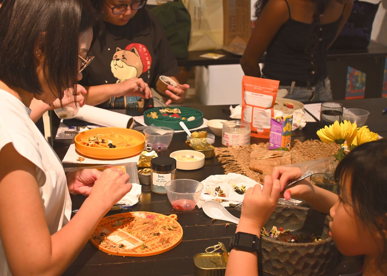 People gathered around a table doing arts and crafts with decorative items, paints, and sunflowers in a dimly lit room.