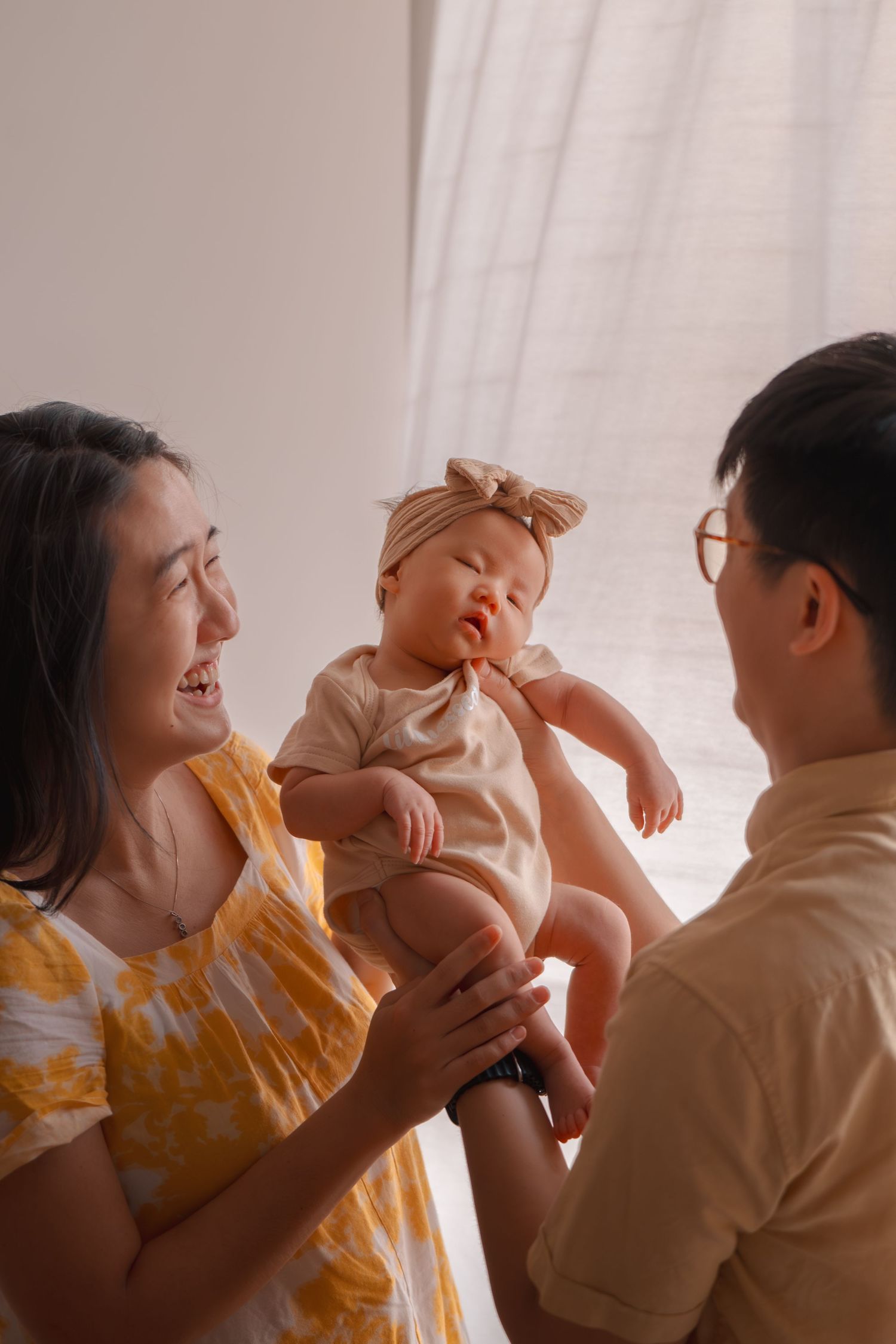 Happy Asian parents smiling and holding their adorable baby girl in a bright, warmly lit room.