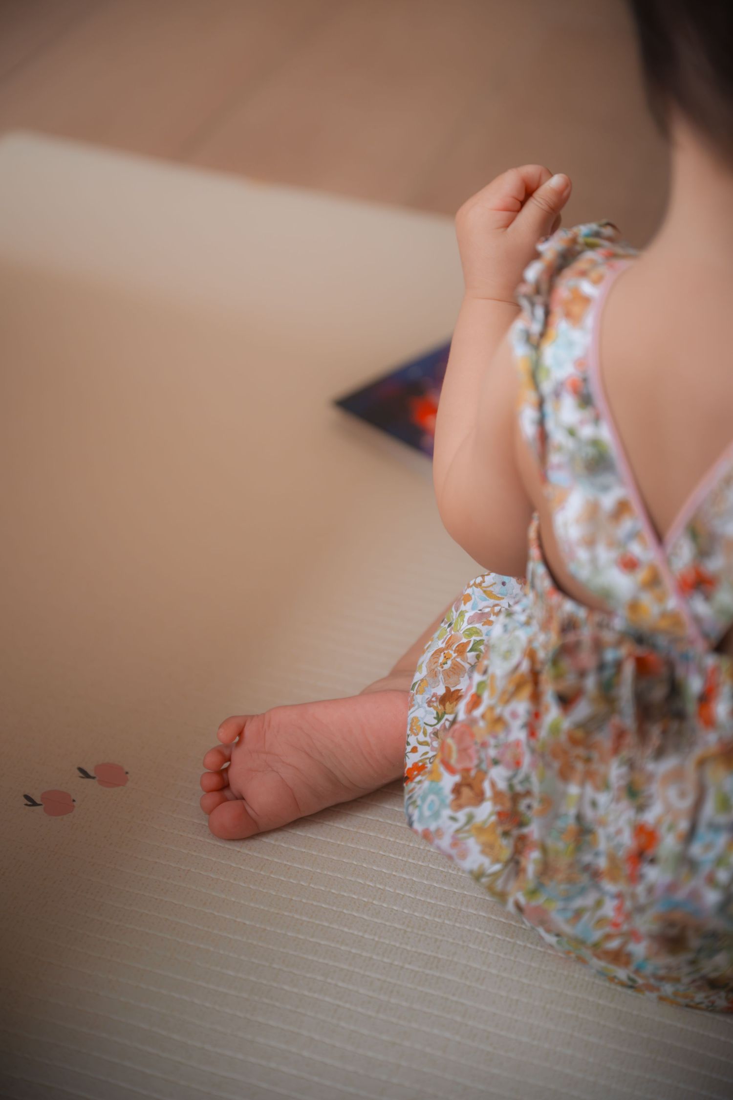 A baby girl in a floral dress sits barefoot on a beige mat, reaching for small colorful pieces.