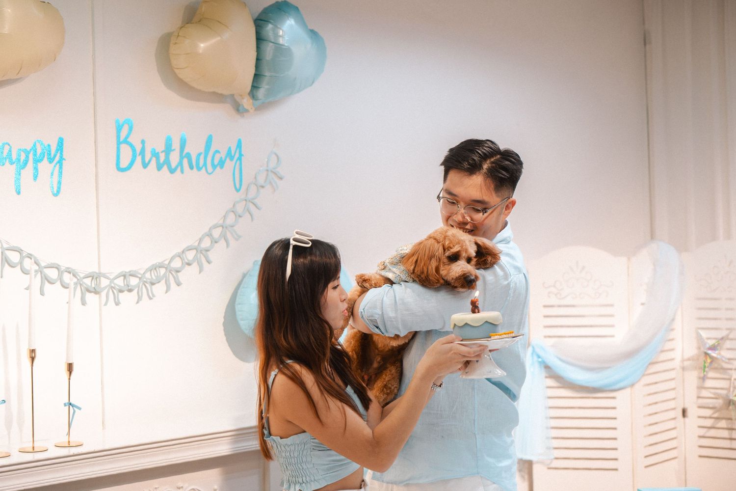 Couple celebrating their dog's birthday party with balloons and Happy Birthday sign in a decorated room.