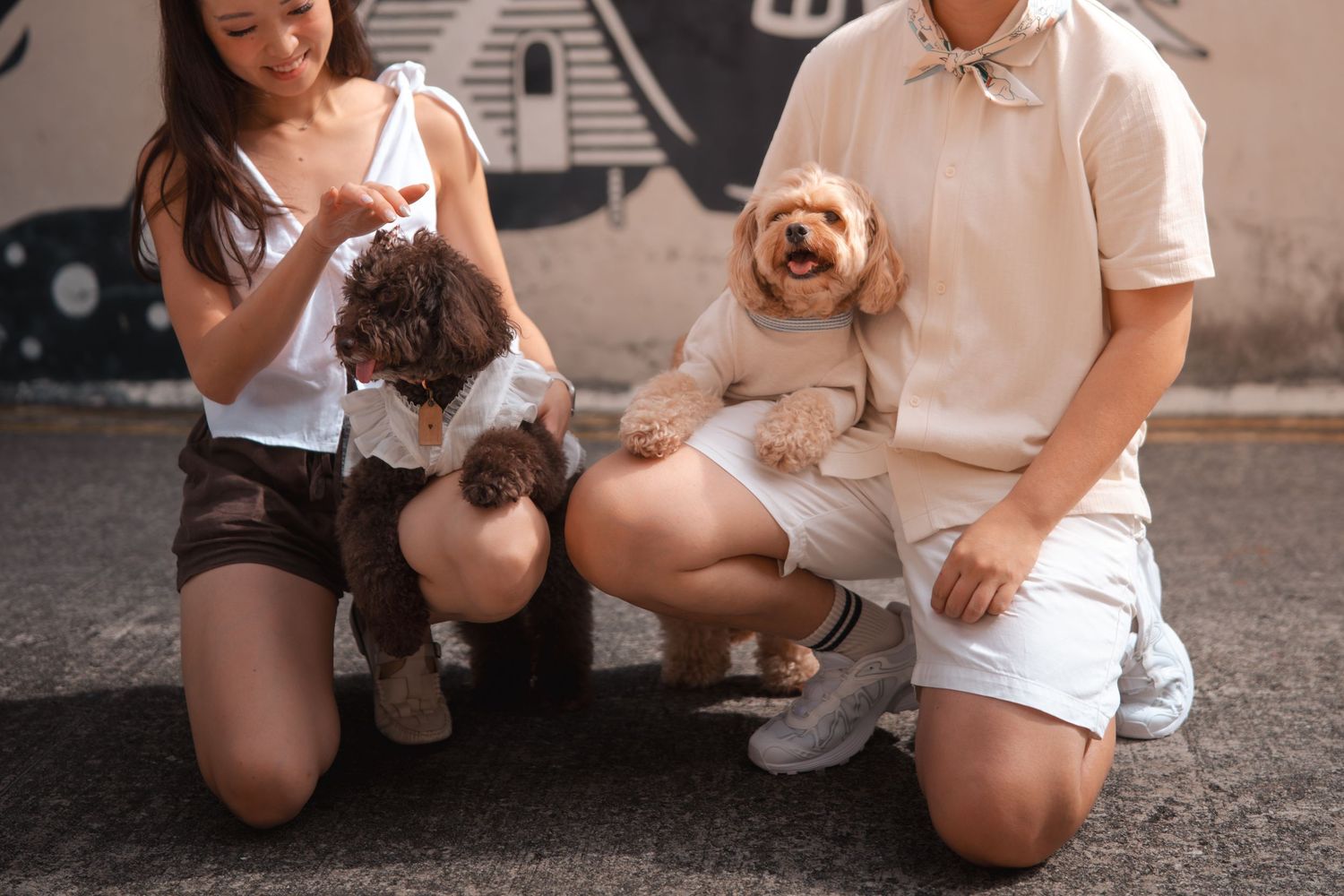 Couple kneeling outdoors holding two small fluffy dogs, dressed in coordinating neutral outfits near a mural wall.