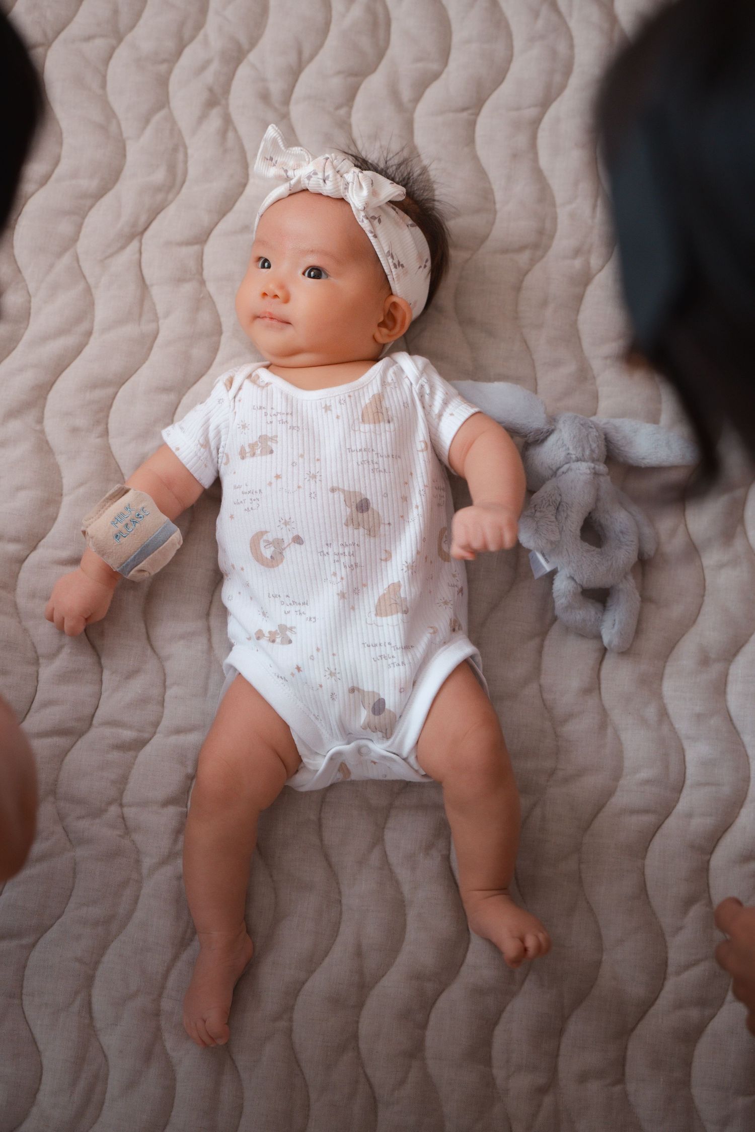 Baby wearing white floral onesie and headband lies on textured grey blanket in multiple poses.