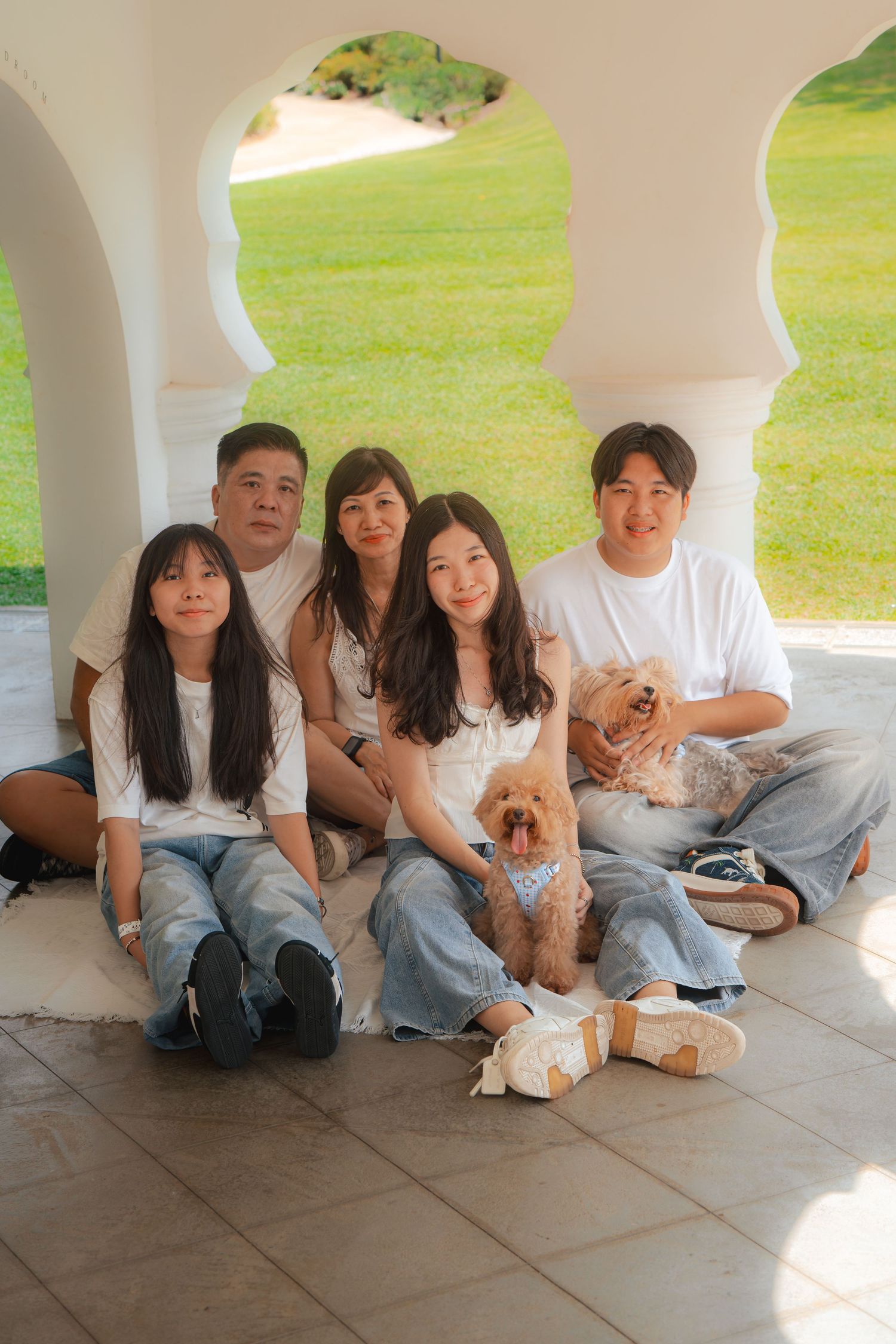 Asian family of four with a dog posing under white arches on a tiled patio with green lawn background.