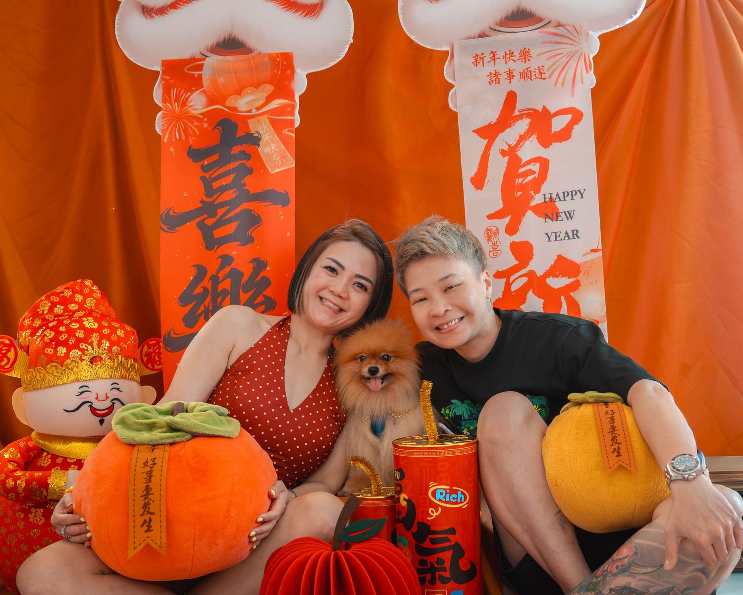 Two people pose with a Pomeranian dog amid vibrant Chinese New Year decorations including orange lanterns and festive ornaments.