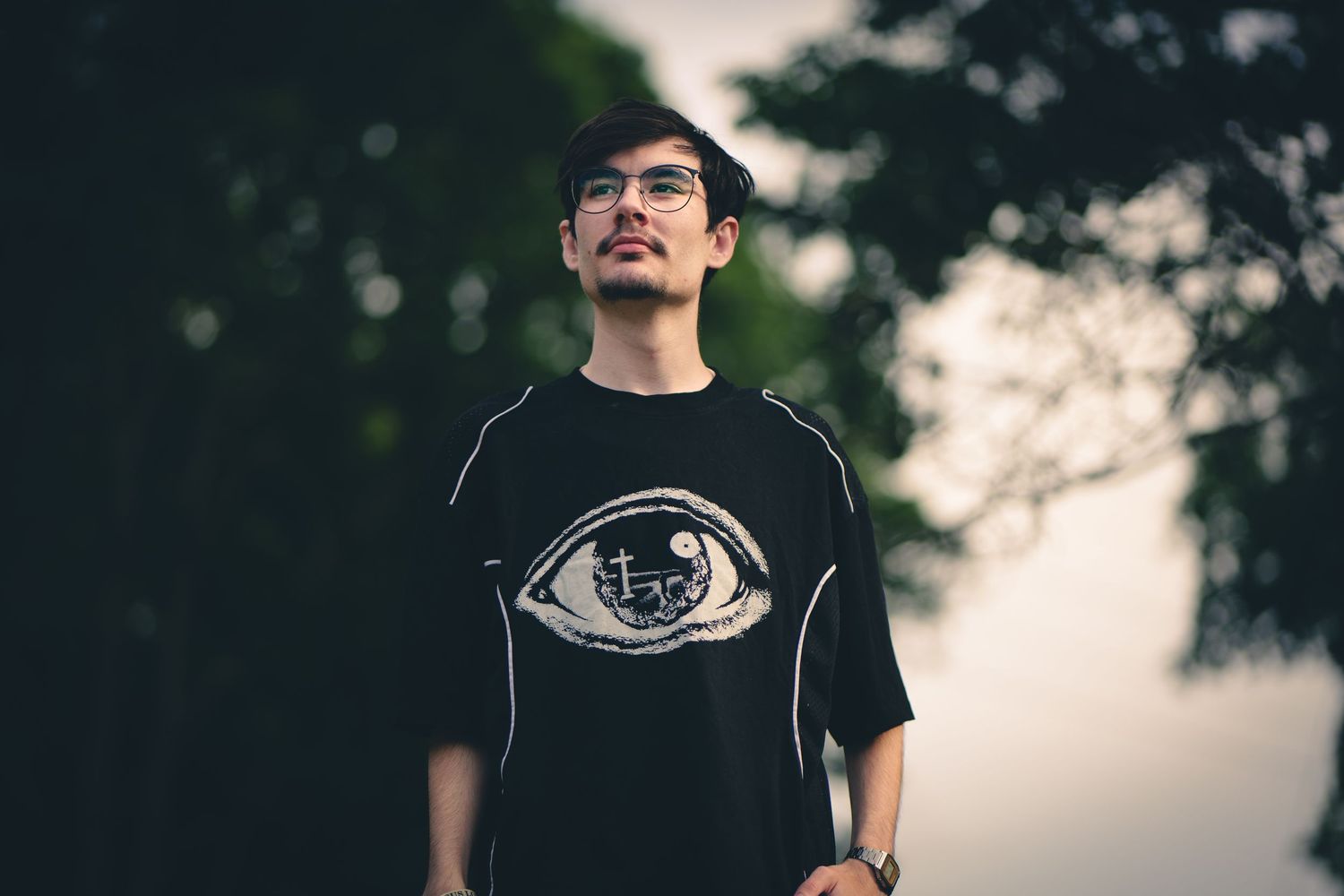 Young Asian man wearing a black graphic eye-print t-shirt, standing outdoors with trees in the background.