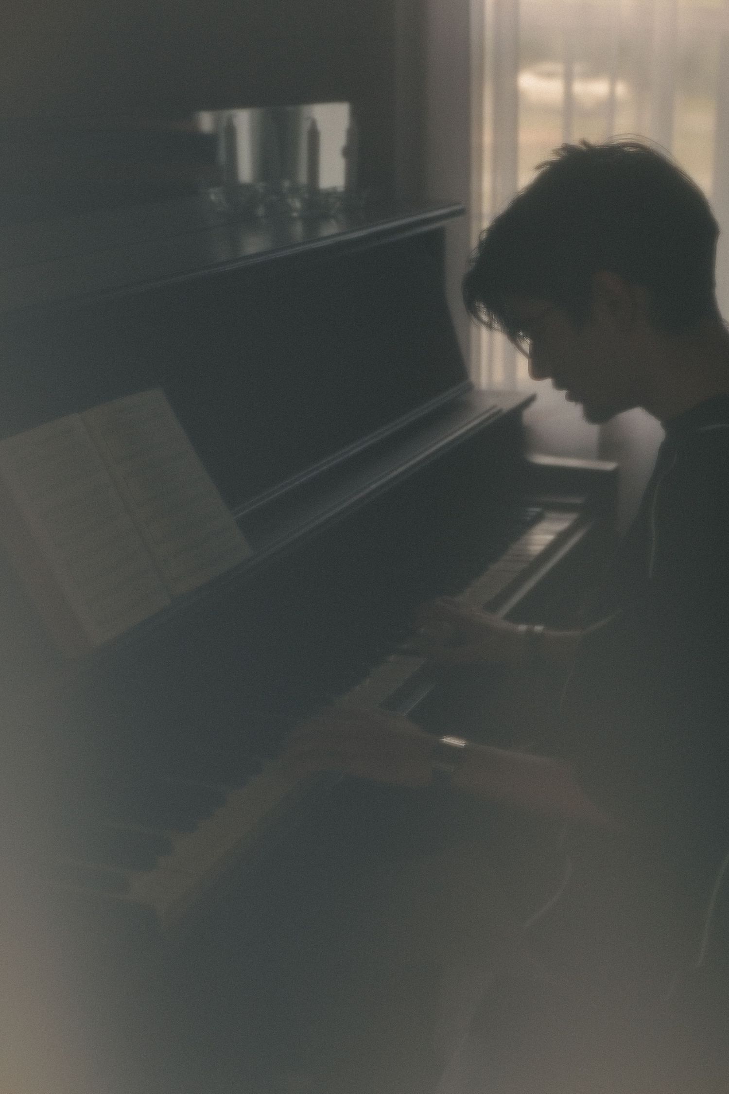 A young man plays an upright piano in a dimly lit room, reading sheet music in a moody, atmospheric setting.