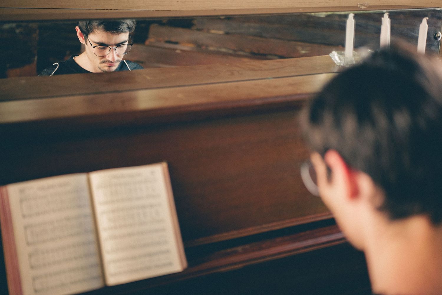 Young man playing piano reflected in mirror with sheet music open, captured in moody indoor lighting.
