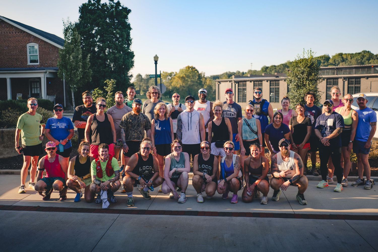 Large group of runners posing together outdoors near a brick building on a sunny day.